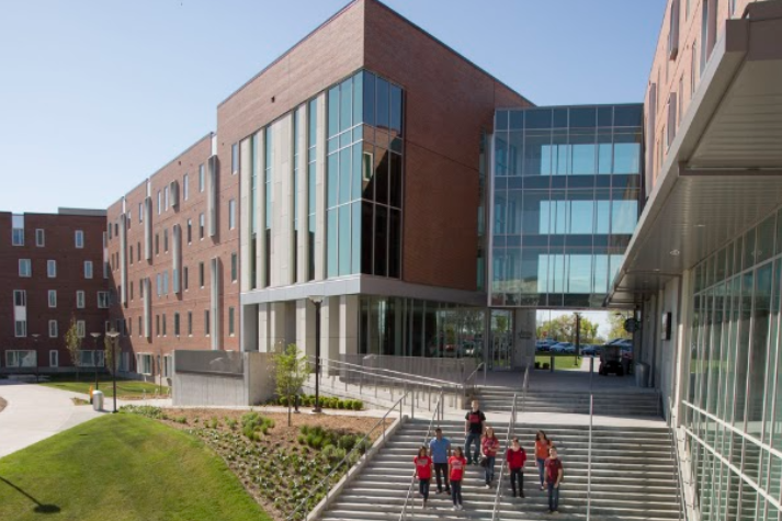 Exterior view of The Crossing, featuring a modern brick-and-glass entrance, wide staircase and accessible ramp, landscaped grounds, and a group of students walking down the steps