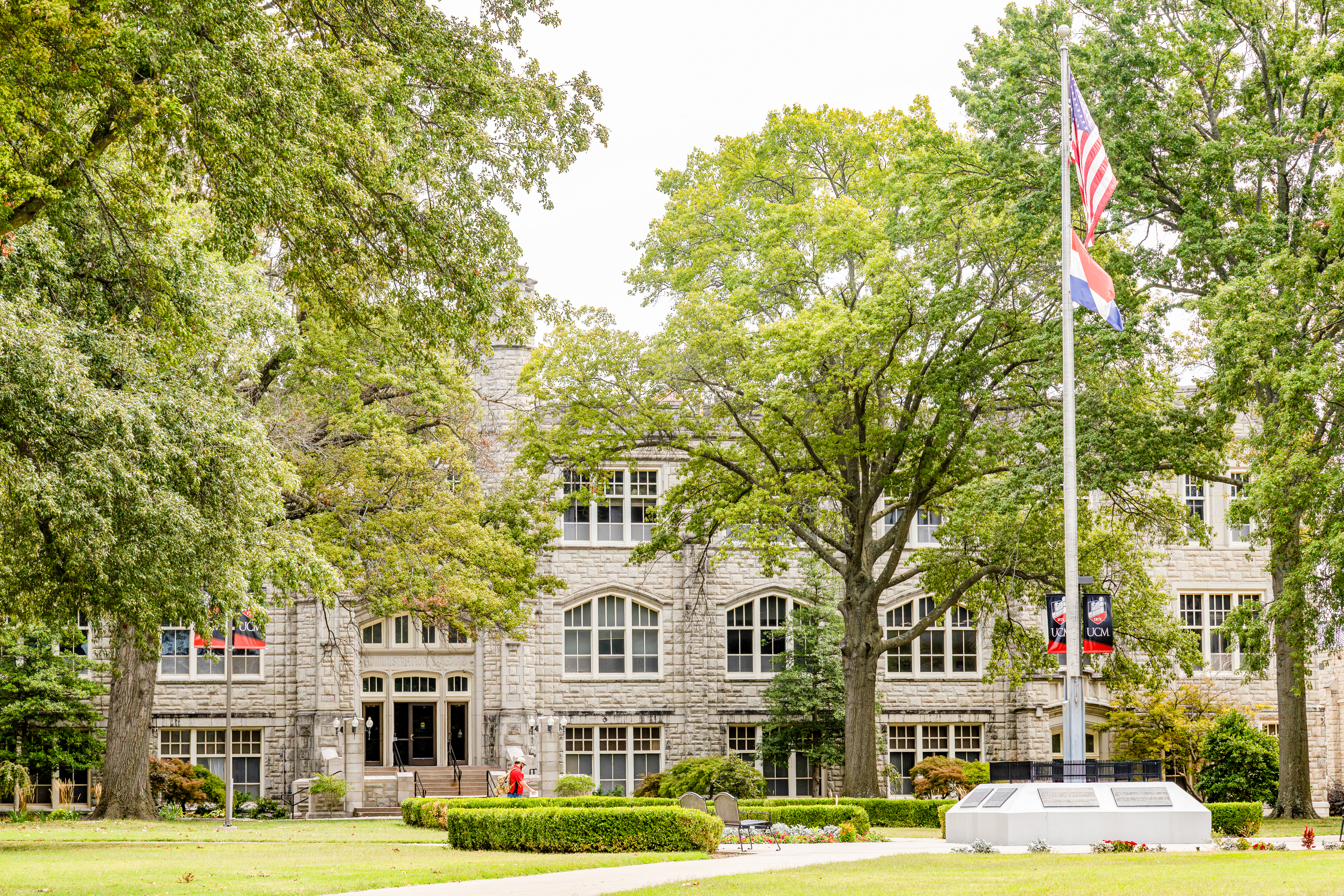 The gray limestone Administration Building is seen with large trees in front of it. A person wearing a red shirt is seen walking past the front of the building, and appears very small in the photo.
