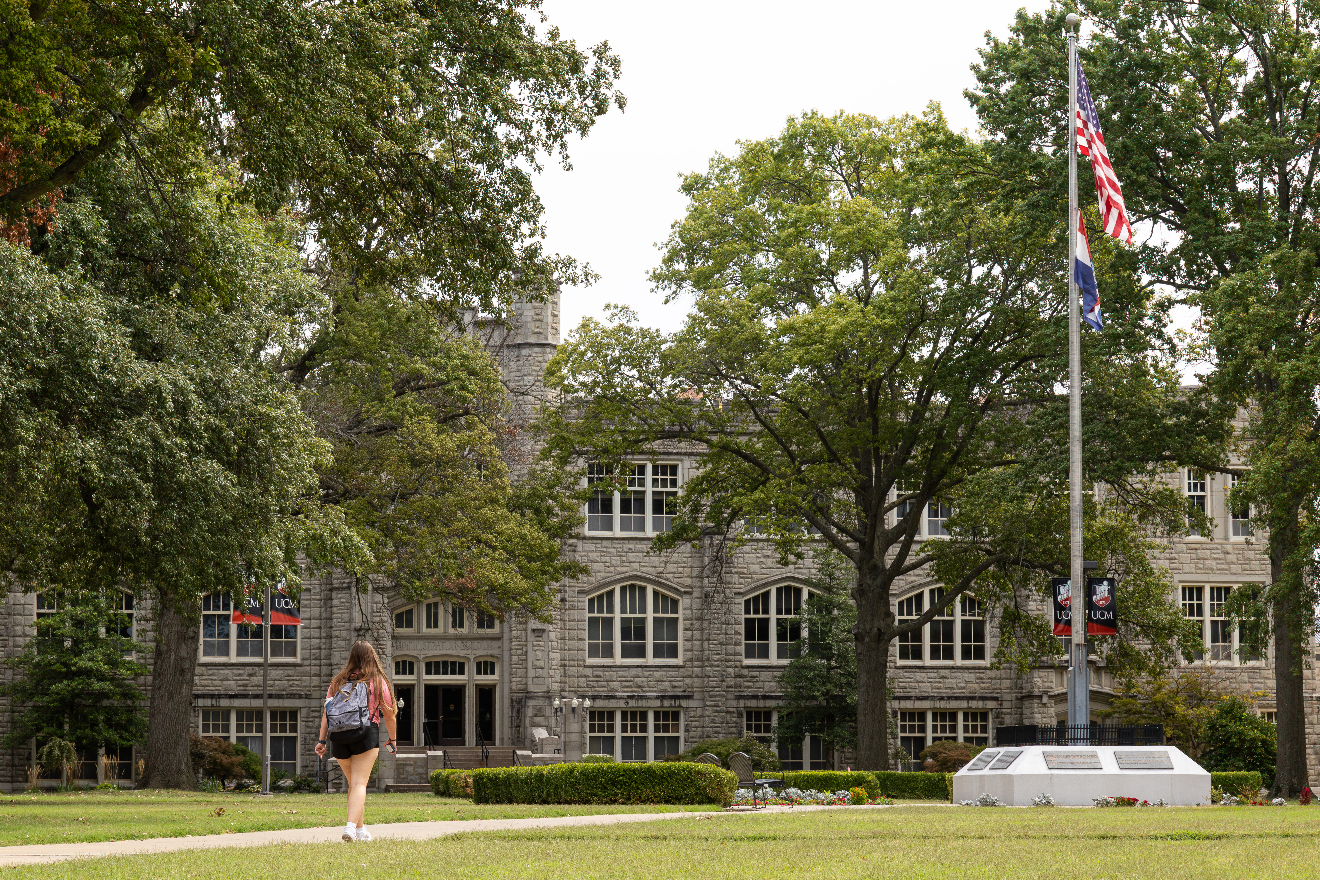 A student wearing a backpack is walking on a sidewalk toward the Administration Building at UCM. A flagpole with the Missouri flag and American flag are on the right. There are several trees in front of the building, which is gray limestone.
