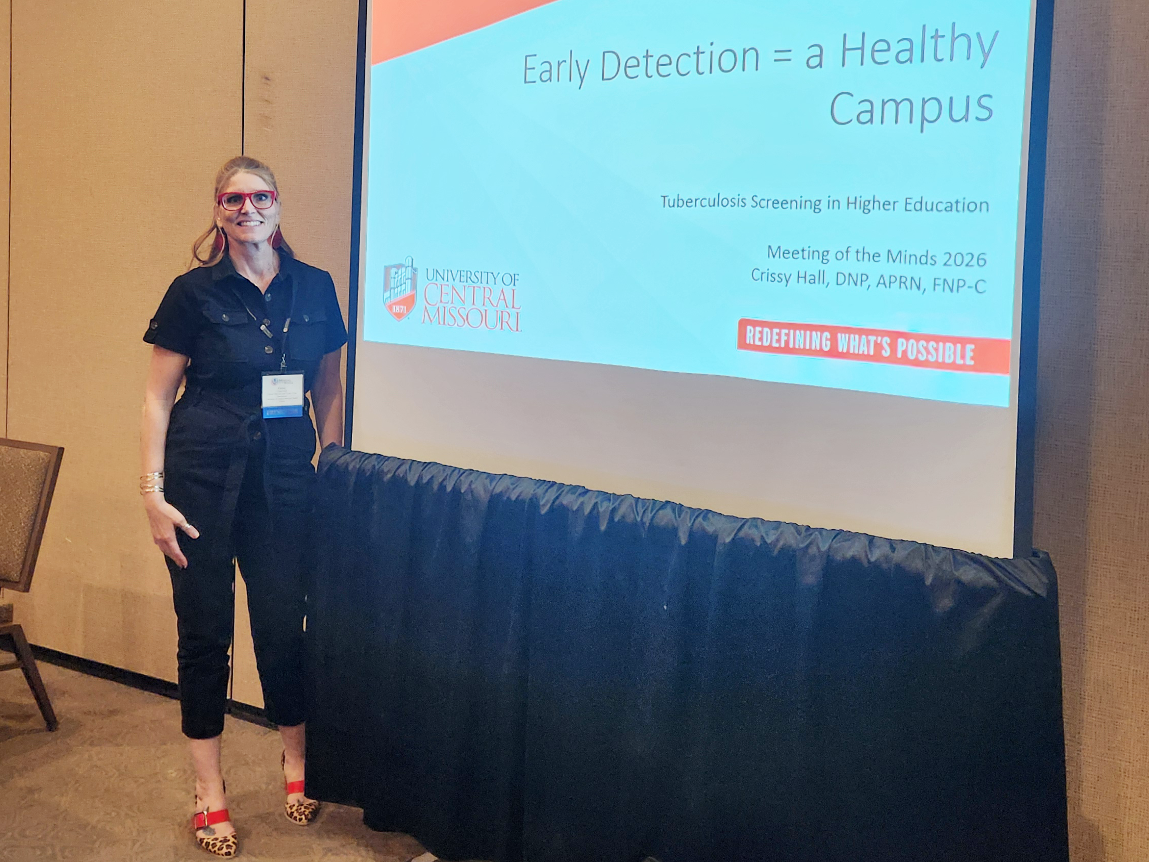 A person wearing a black shirt and black pants stands next to a projection screen that is showing a slideshow presentation titled "Early Detection = a Healthy Campus."