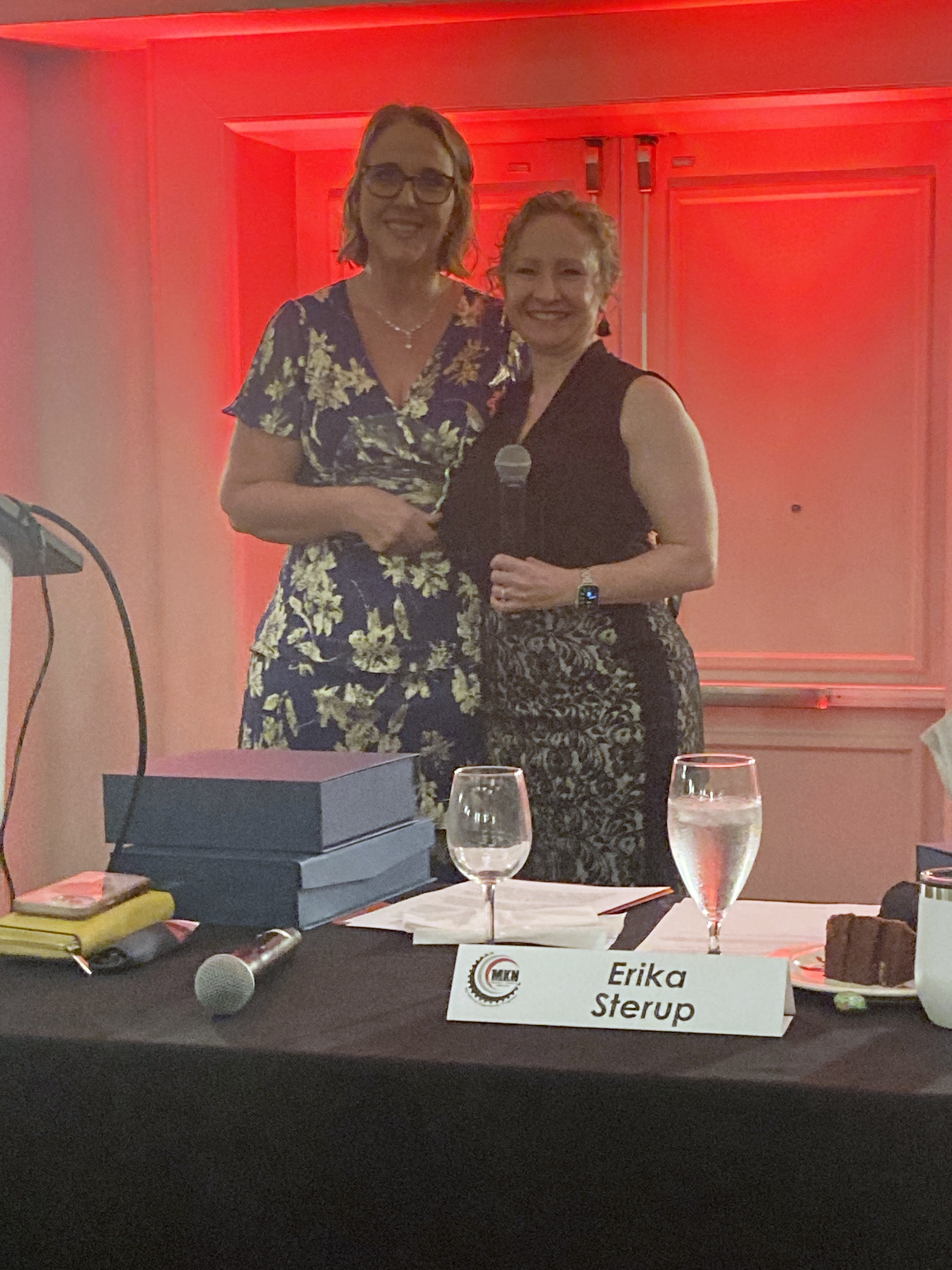 Two people stand next to each other with a table in front of them. The one on the left is wearing a blue floral dress and holding an award plaque. The one on the right is wearing a black dress and holding a microphone.