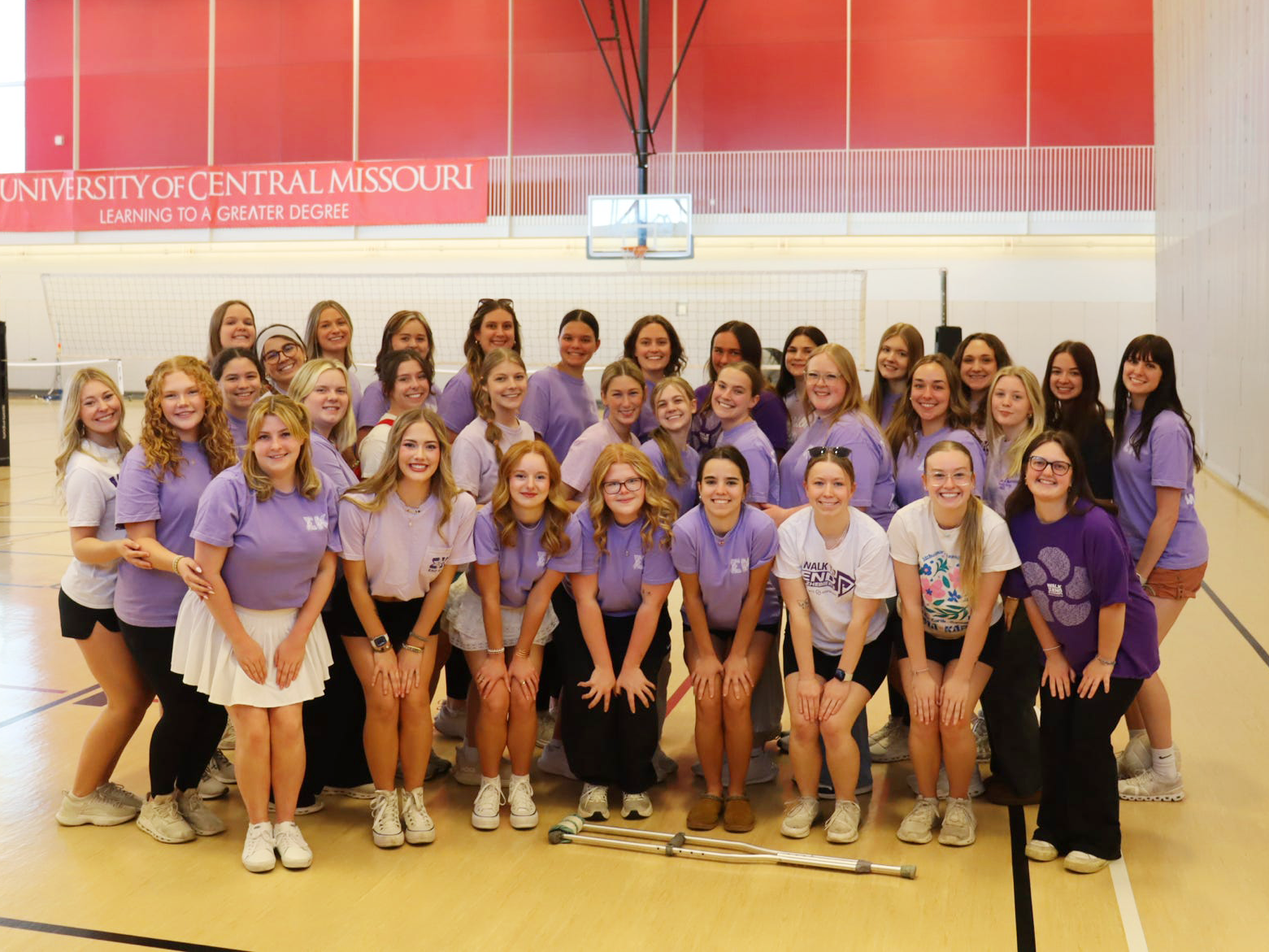 A group of more than 30 sorority members stand together in the middle of a volleyball court. Almost everyone is wearing a purple T-shirt, with a few wearing a white T-shirt.