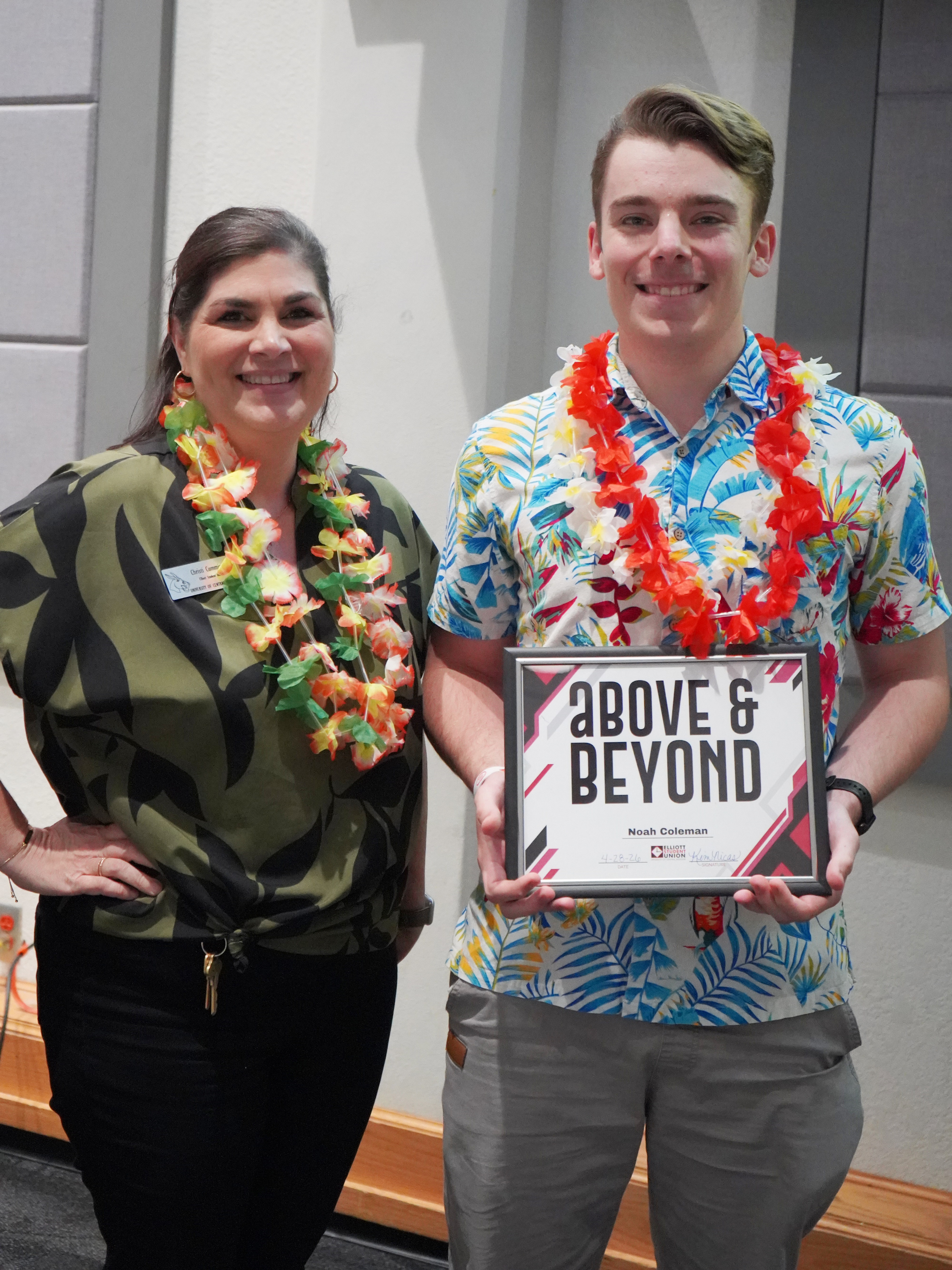Two people stand next to each other in a conference room in the Elliott Student Union. Both are wearing Hawaiian-themed shirts and flower leis. The person on the right is holding an award certificate.