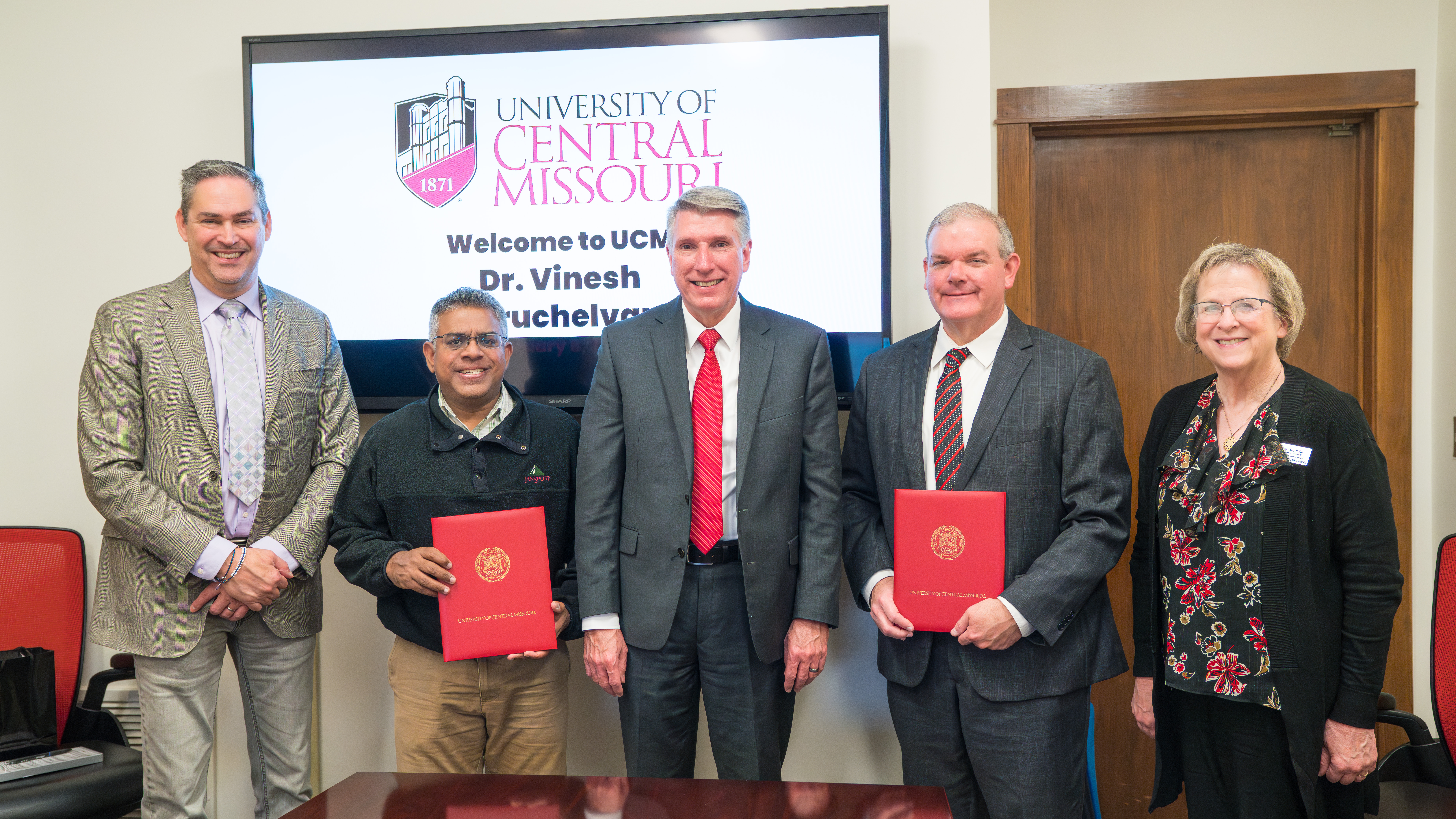 UCM and APU leadership Five people stand in a row in a conference room. Two of them are holding red UCM folders.