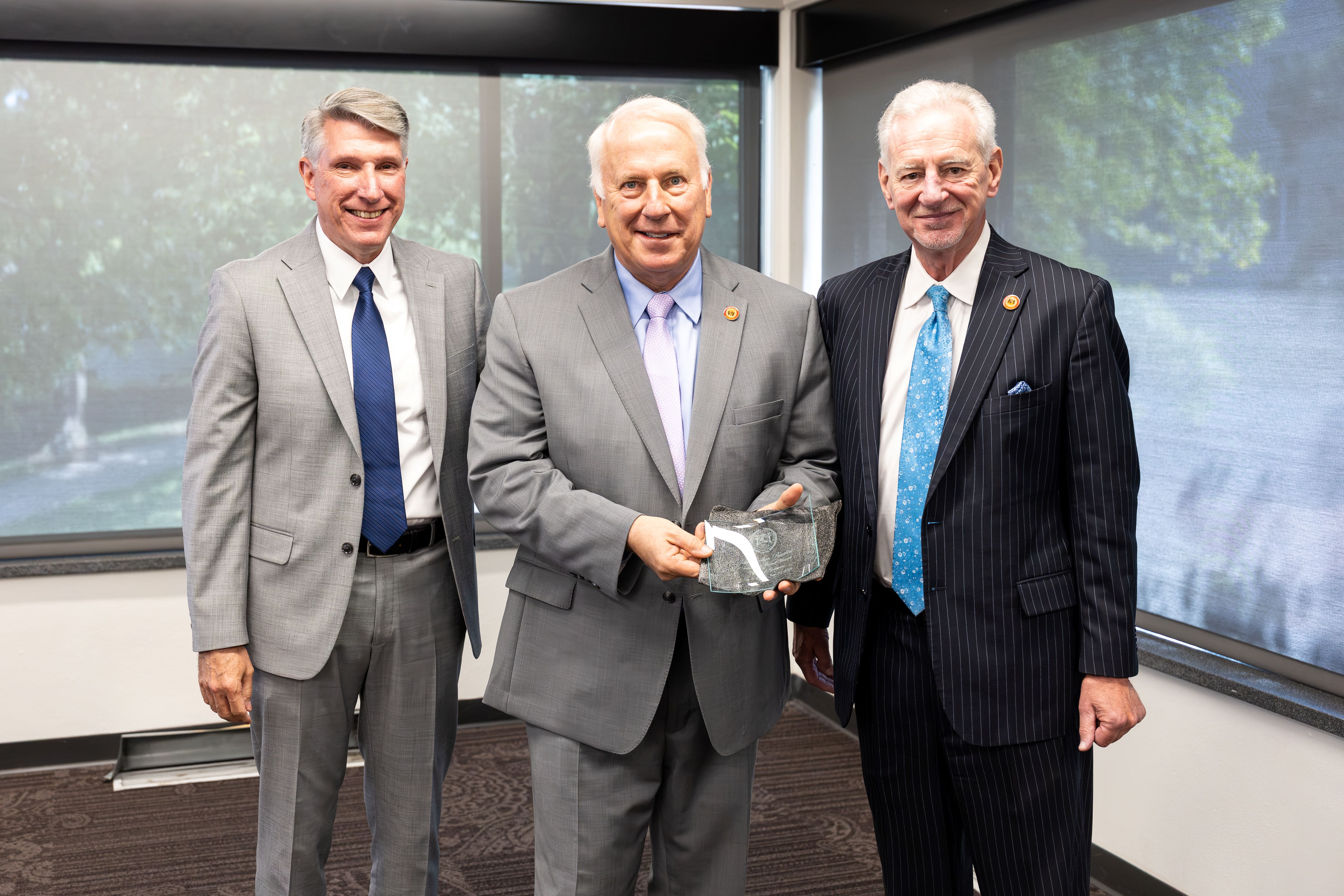Incoming & outgoing board presidents Three people wearing suits stand next to each other in a meeting room with large windows behind them that have a view of the UCM campus. The center person is holding a recognition plaque.