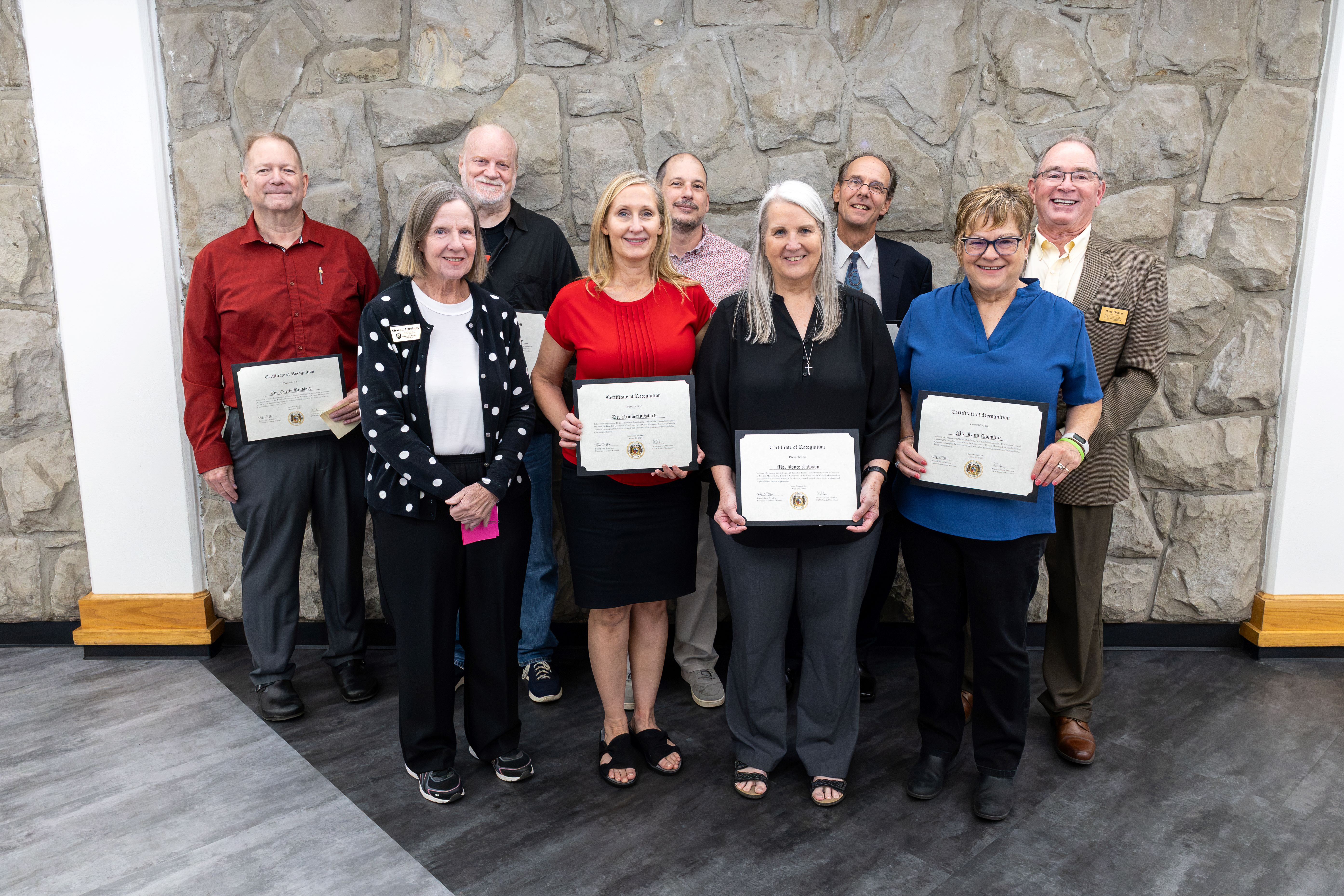 New emeriti faculty and staff members Four adults stand in the front row and five stand in the back row. They are smiling and looking at the camera while standing in a hallway in front of a gray stone wall. Several people are holding certificates.