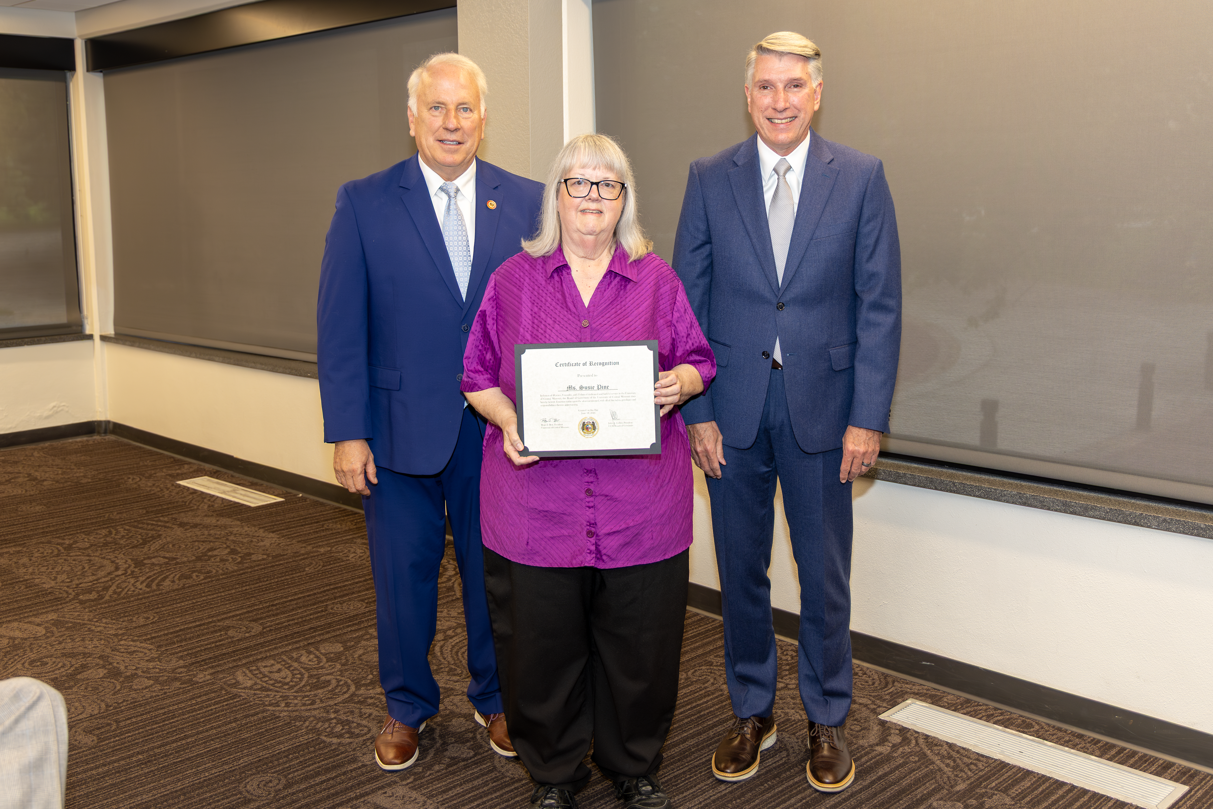 Three people are standing next to each other in a meeting room in the Elliott Student Union - Board President John Collier, retiring employee Susie Pine and UCM President Roger Best. The person in the middle, Susie, is wearing a purple button-down shirt, black pants and glasses and holding an award certificate. John is on the left and Roger is on the right and both are wearing a blue suit. 