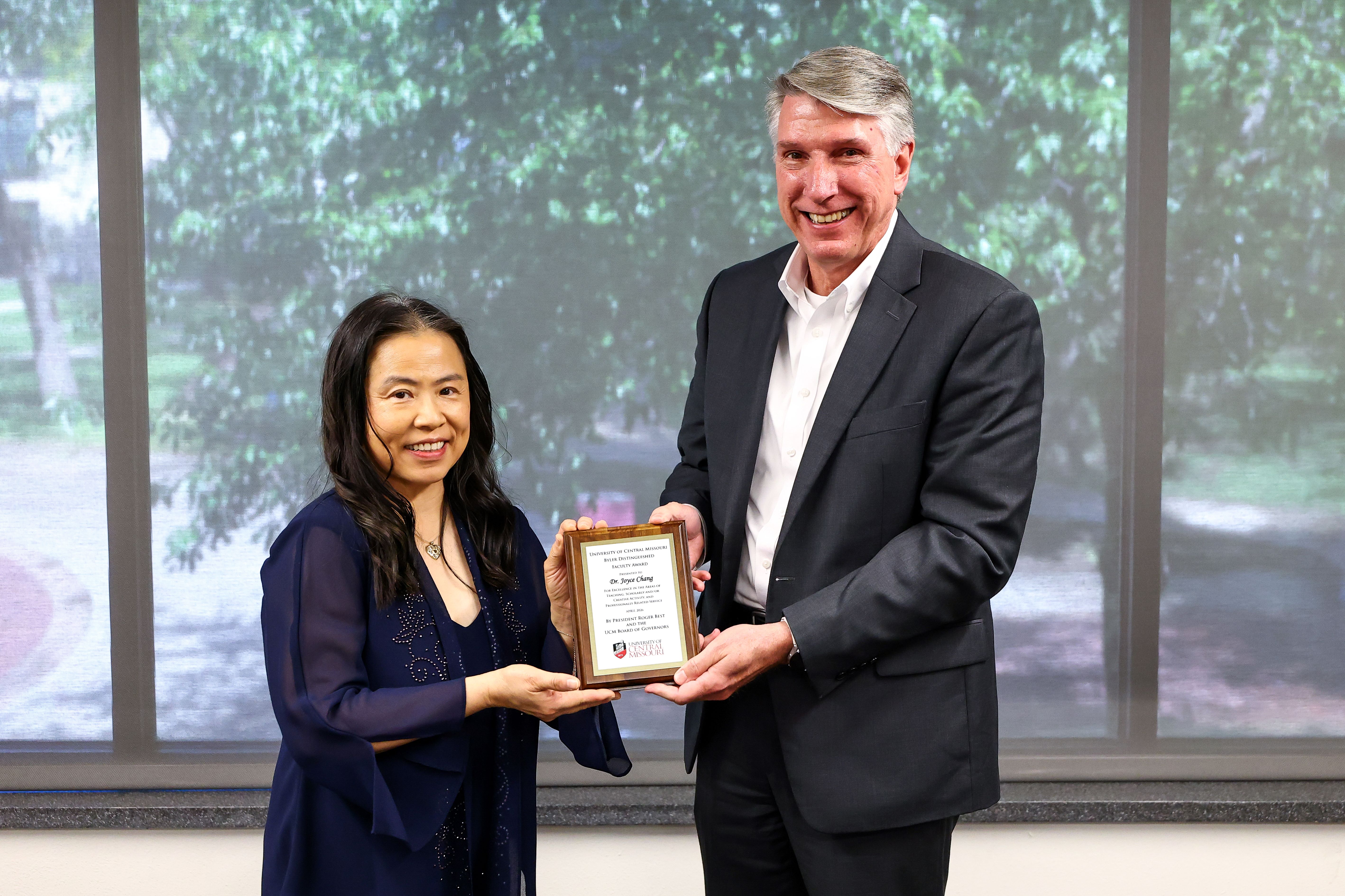 A woman stands on the left and a man stands on the right. They are each holding one side of the Byler Distinguished Faculty Award plaque. They are standing in a room in the Elliott Student Union with a window behind them.