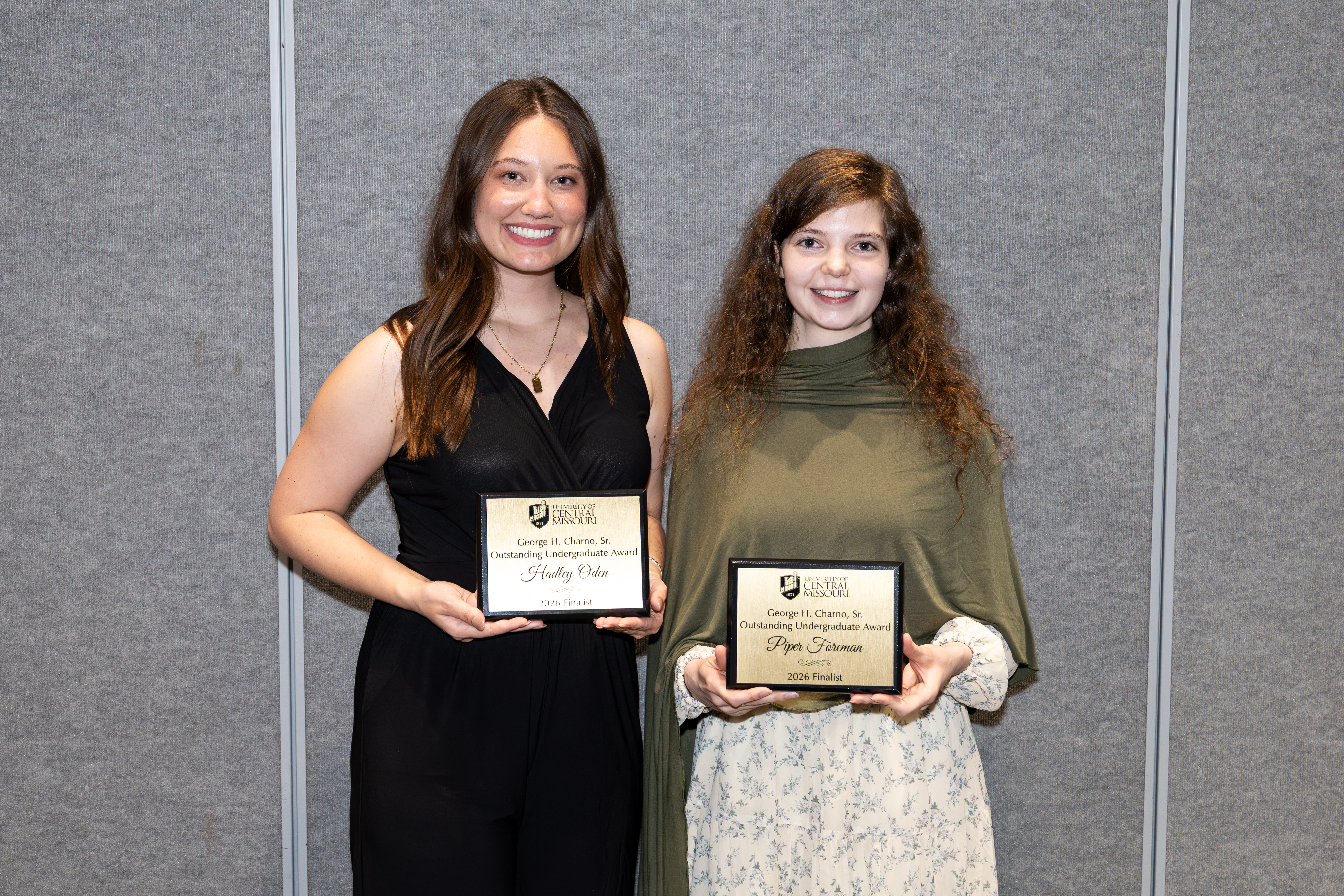 Two people stand in front of a gray wall and both are smiling and holding gold award plaques
