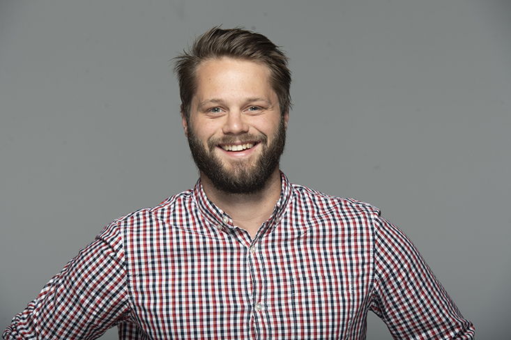 A man wearing a plaid shirt is smiling in front of a gray background