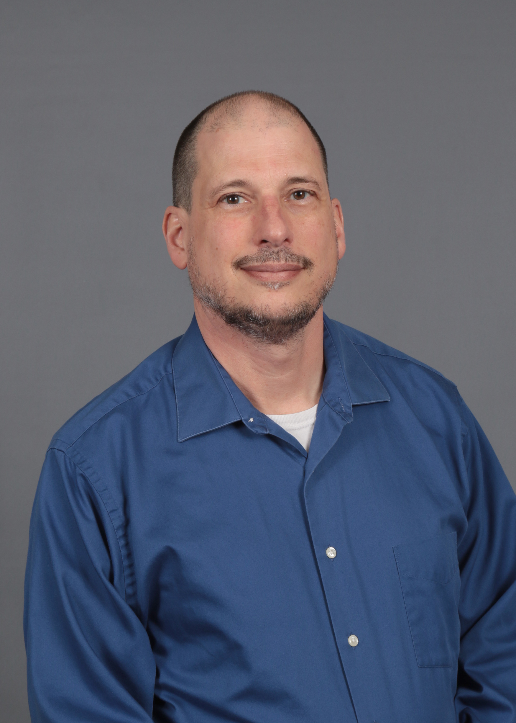 A person is seen from the shoulders up in a professional portrait with a gray background. They are wearing a blue collared-shirt.
