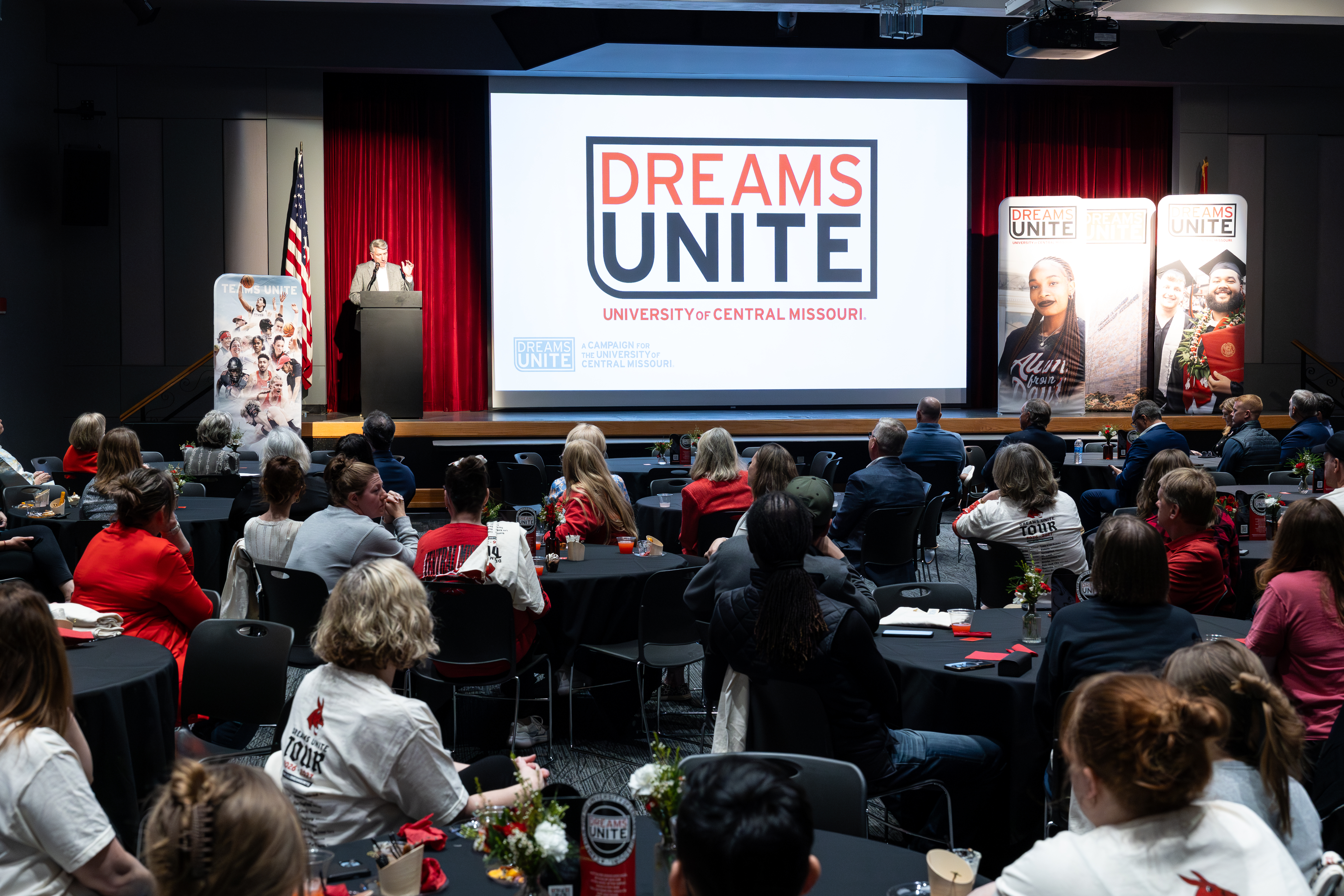 A person wearing a gray suit stands behind a podium on a stage and talks to the audience. Next to the person is a large screen showing the Dreams Unite campaign logo. The event is in the large Elliott Student Union ballroom and people are sitting in chairs at round black tables facing the stage.
