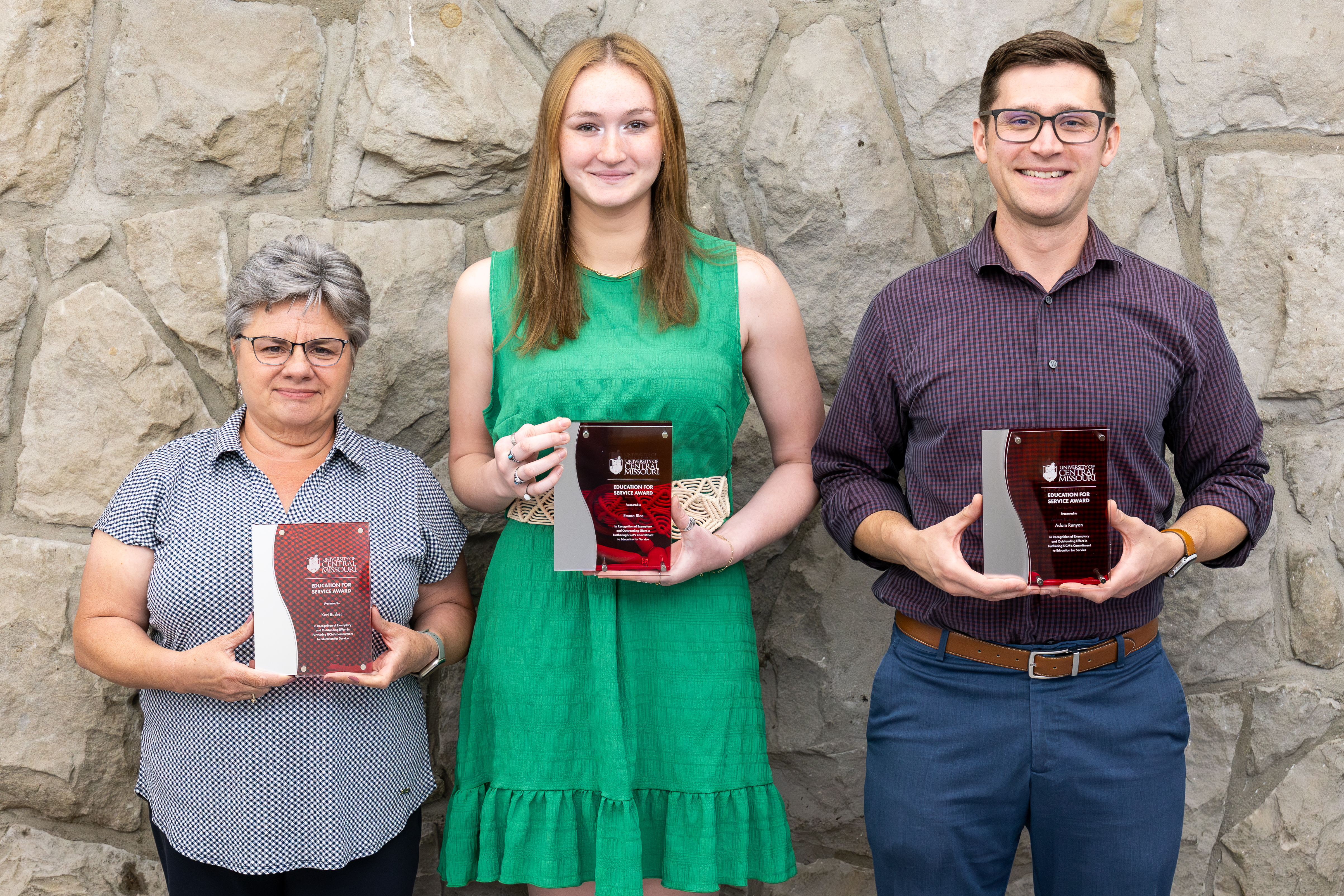 Three people stand next to each other in front of a gray stone wall. Each person is holding a red and silver acrylic award plaque.