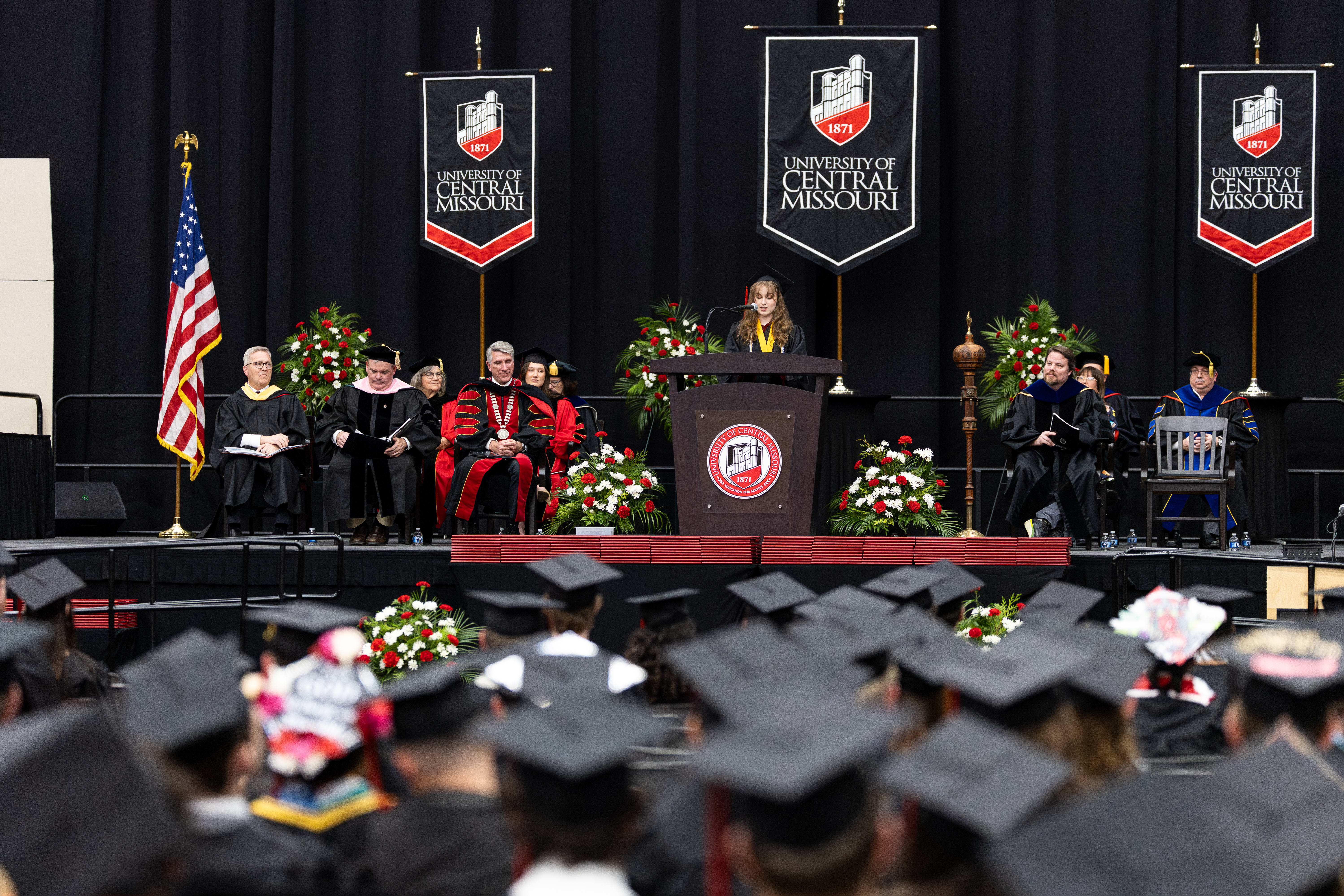 A young woman is standing behind a podium giving a speech. There is a black curtain behind her. She is wearing a black graduation cap and gown. On the stage behind her are several UCM leaders sitting in chairs also wearing graduation regalia. Dozens of graduates are seen in front of her, sitting in chairs on the floor in front of the stage. They are all wearing graduation caps and some are decorated.