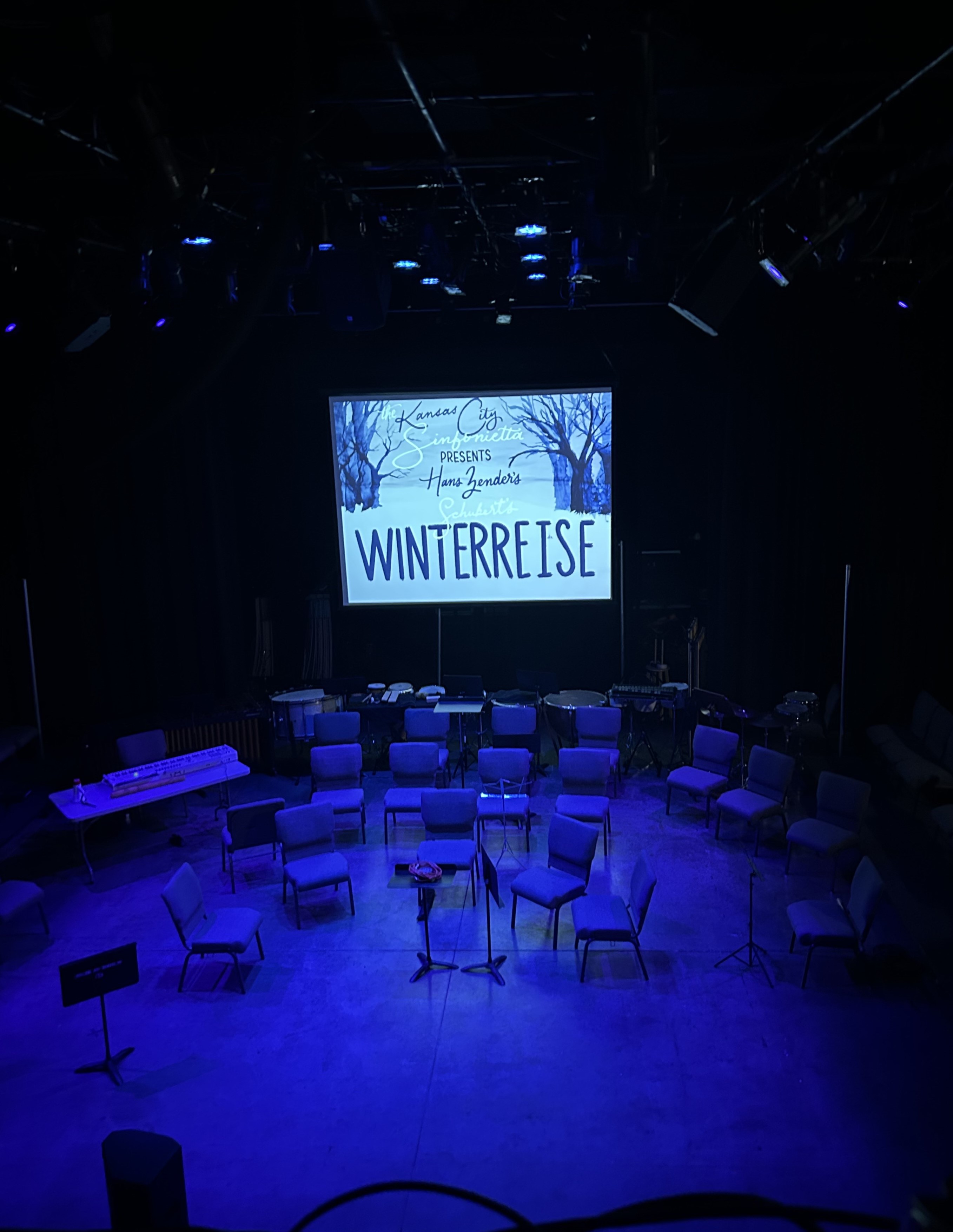 A stage filled with chairs is darkly lit in blue light