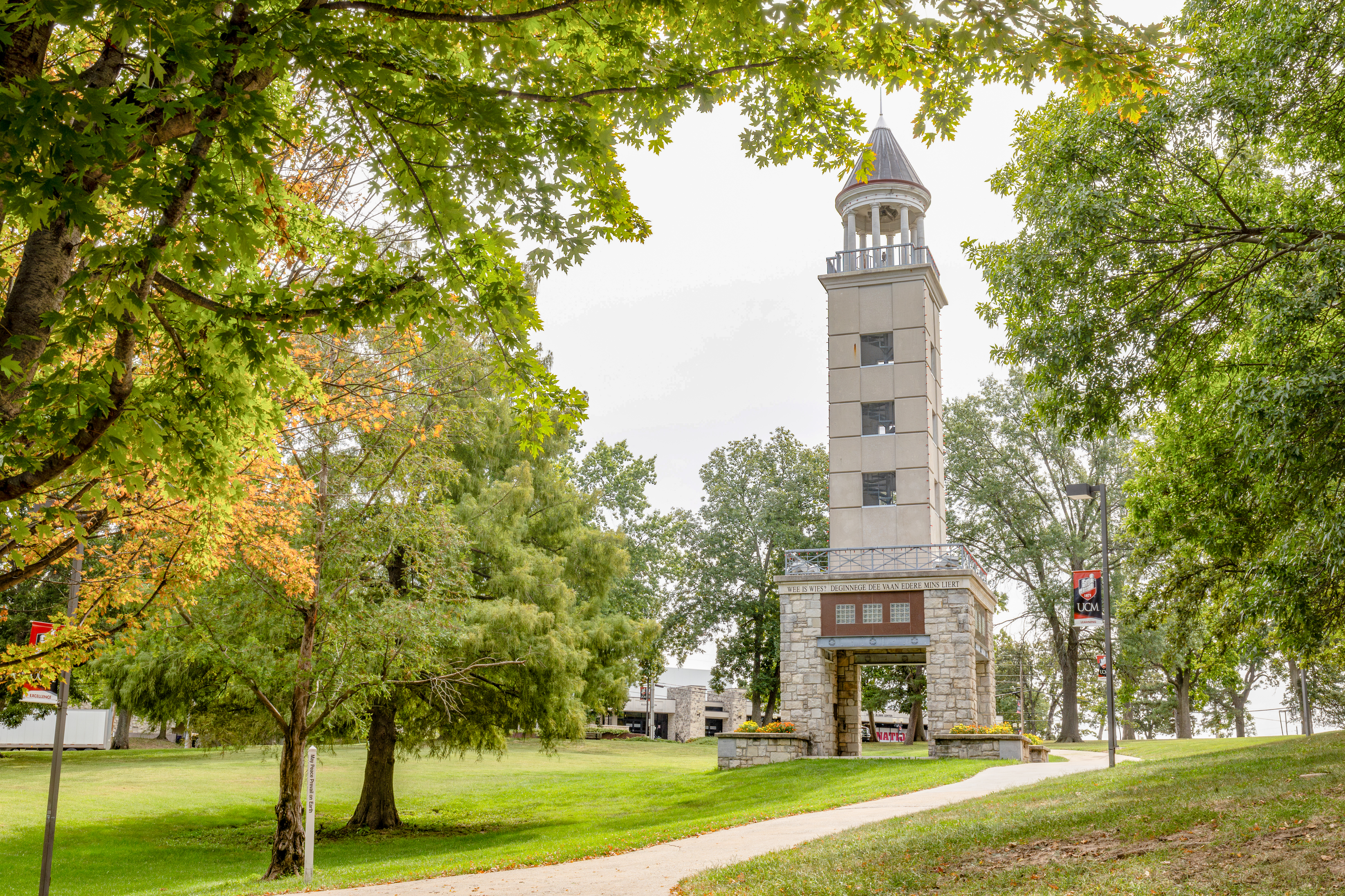 The gray stone Friendship Tower is seen on the right side and several trees, some green and some starting to turn fall colors, are on the left. There are orange and yellow flowers planted in front of the tower and there are UCM banners on the light poles along the sidewalk that passes by the tower.