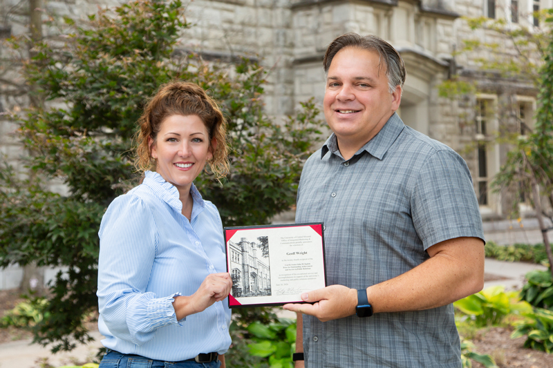 Two people stand in front of a stone building on the UCM campus. Plants and trees are seen behind them. The people are smiling and looking at the camera, and they are each holding one side of an award plaque.