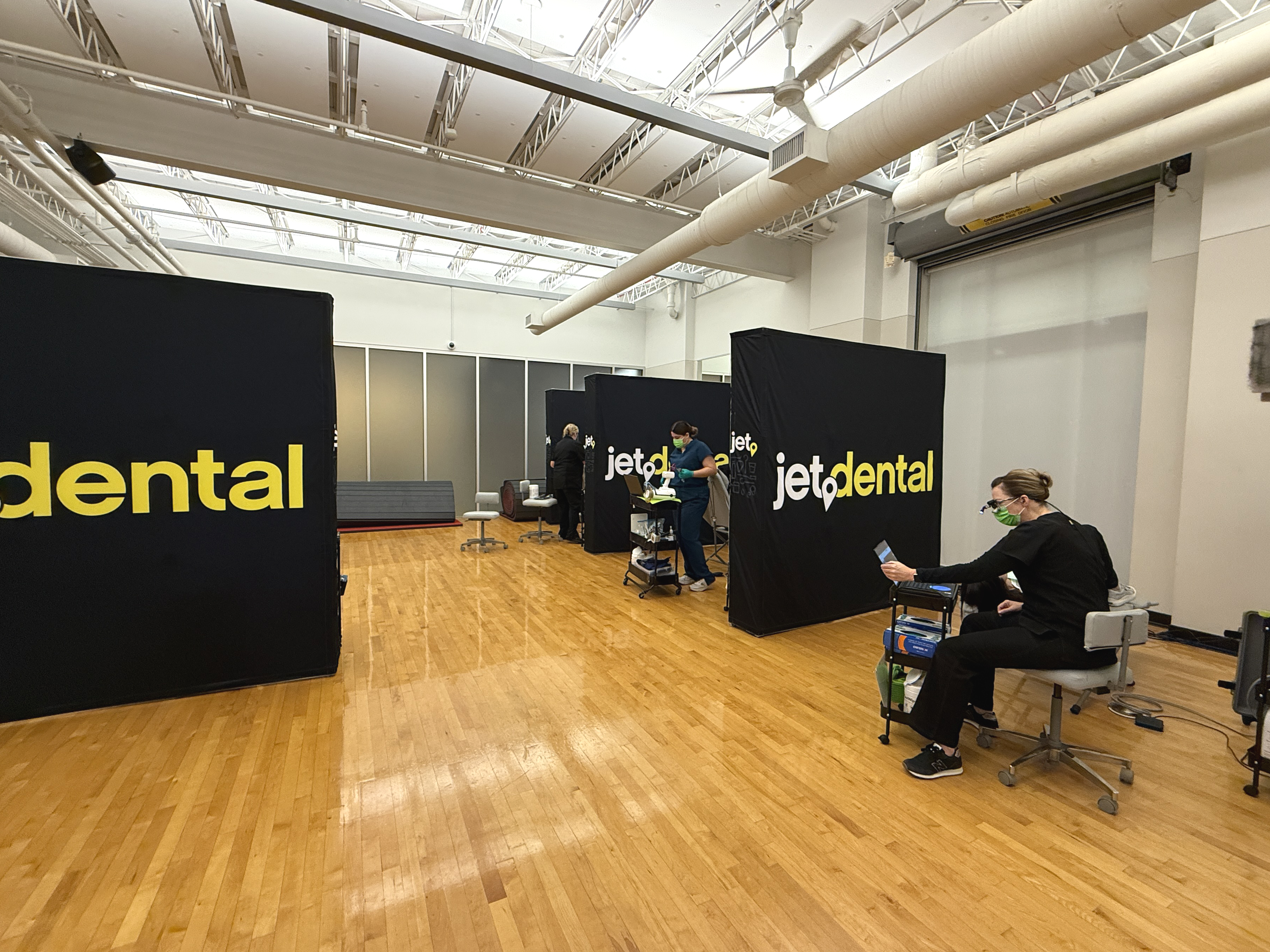 Two dental hygienists work with patients while a third preps for an incoming patient in a room in the Student Recreation and Wellness Center. There are hardwood floors and tall, black dividers that say "Jet Dental" between each hygienist's setup.