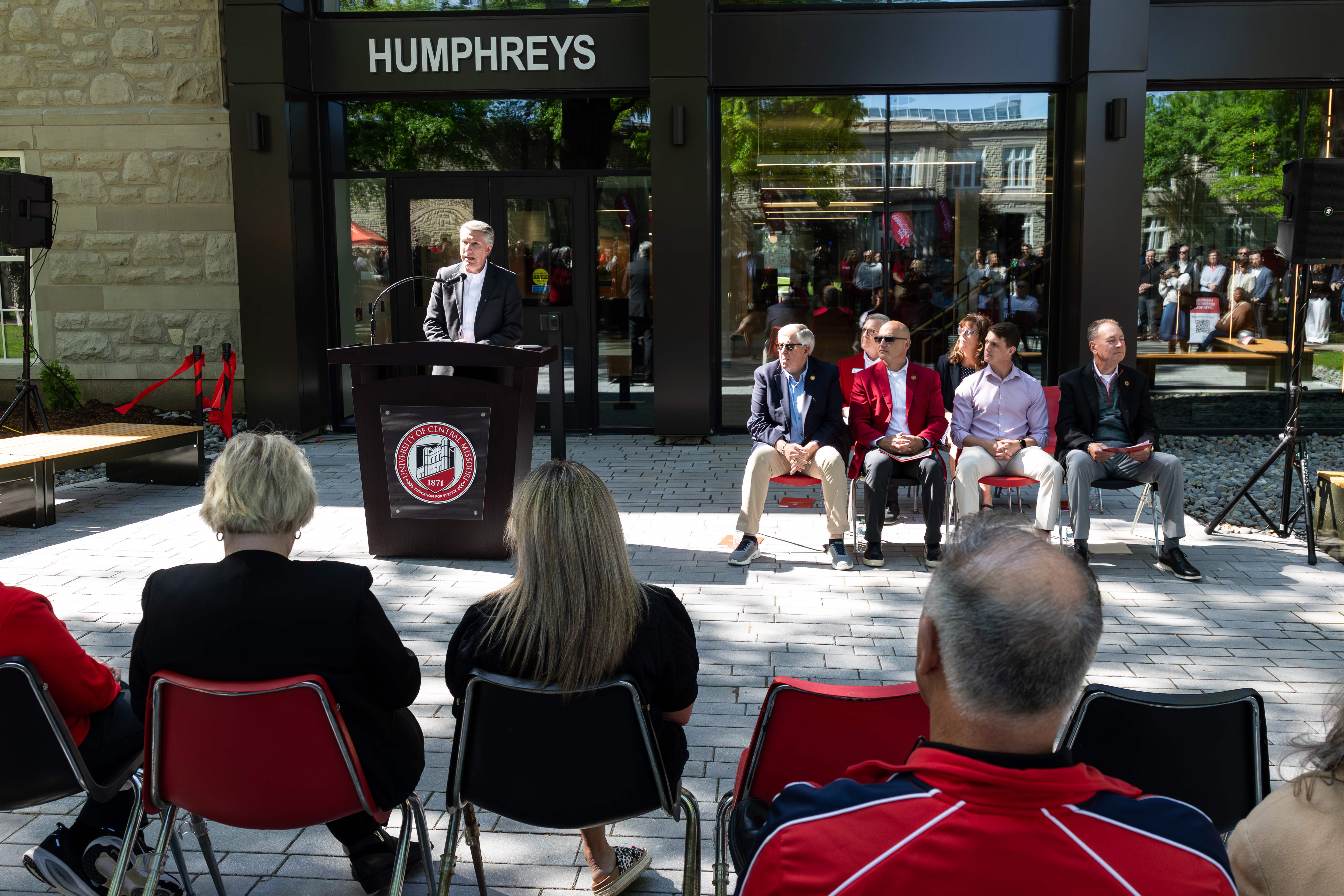 A man stands behind a podium. There are people sitting in two short rows of chairs next to him. The event is outside in front of the Humphreys Building.