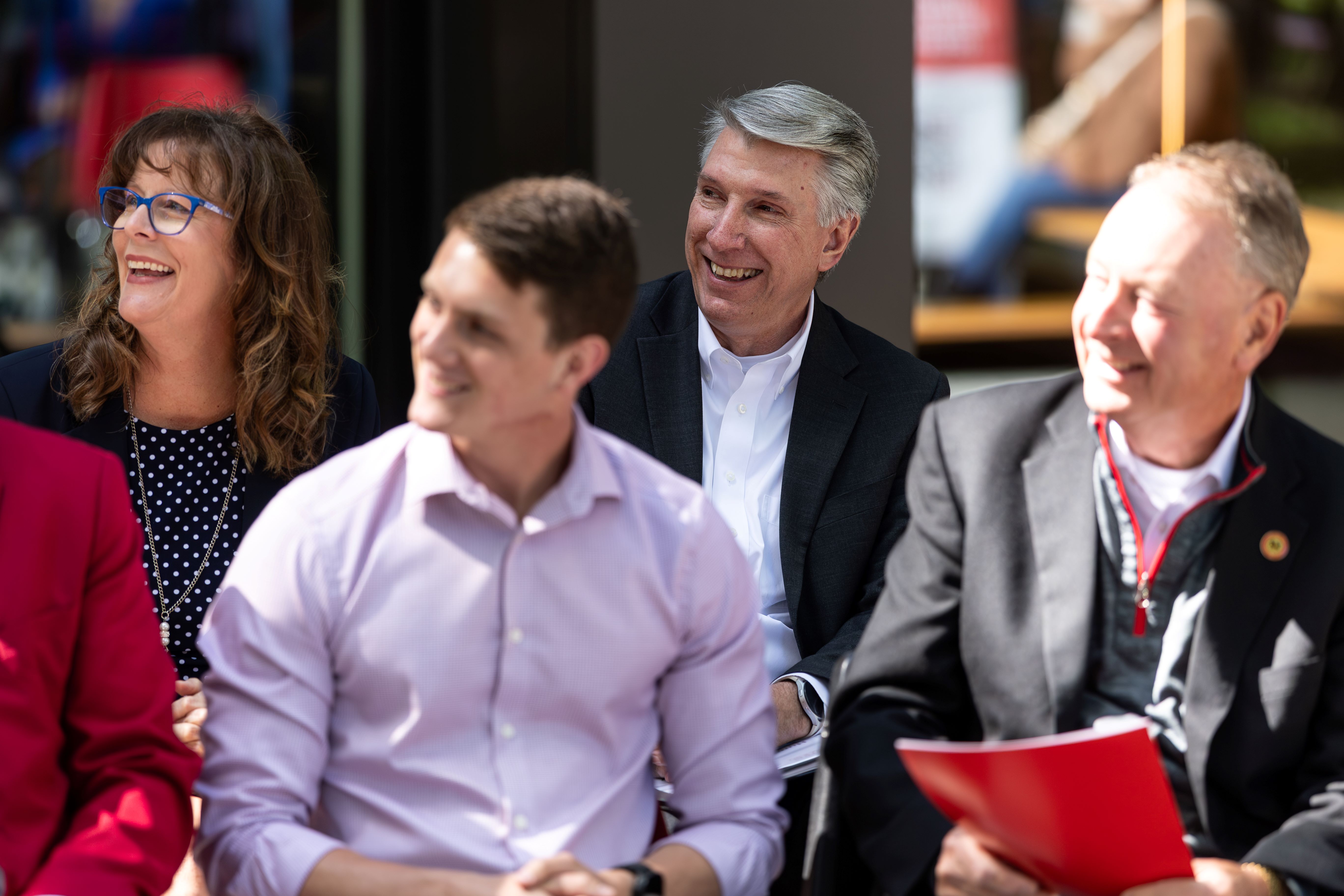 Four people sitting outside in chairs are seen smiling looking to the left toward a speaker who is not in the photo.