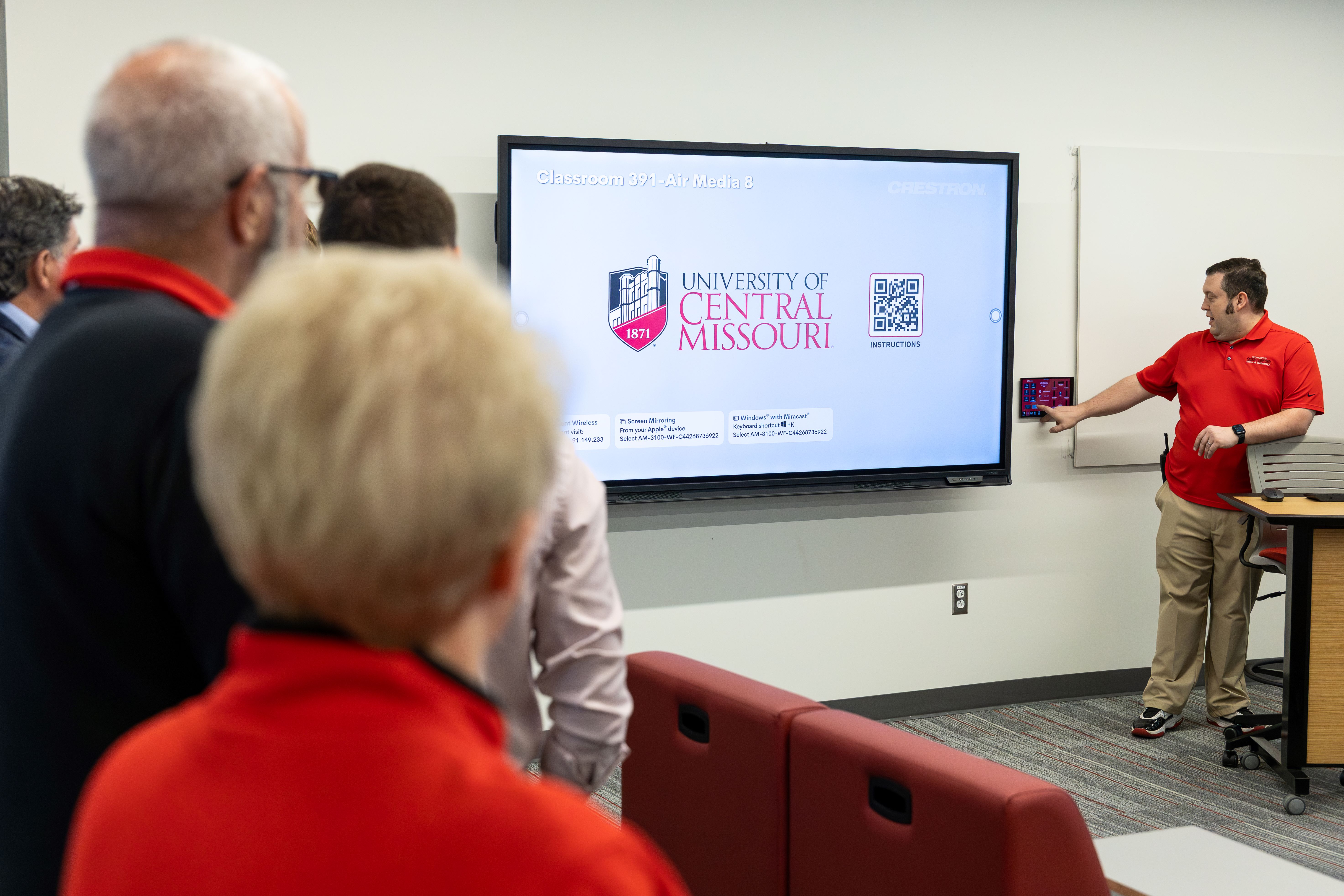 A man in a red polo shirt points to a TV screen as he explains aspects of the classroom to people on a tour.