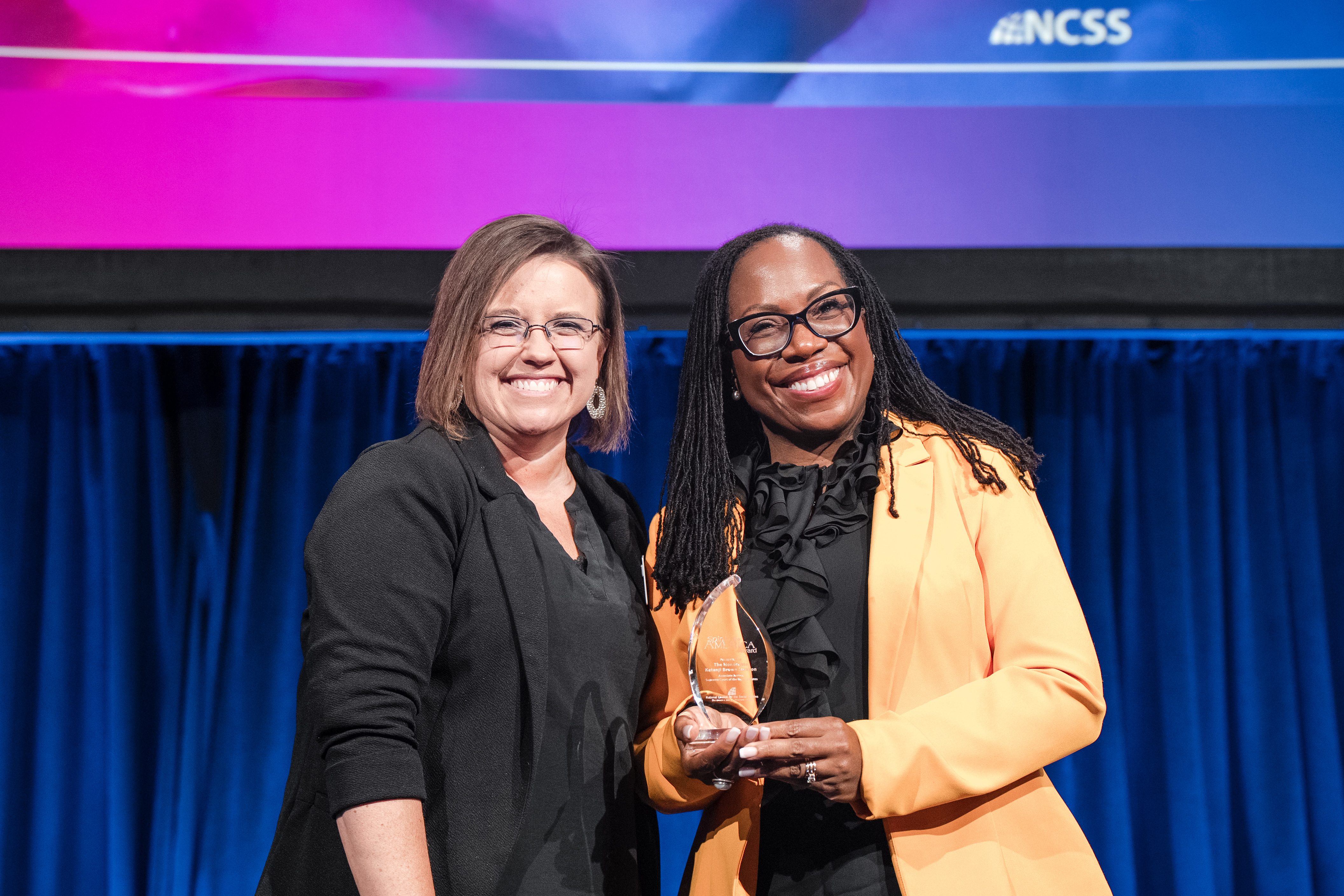 Two people pose for a photo on stage, one is holding an award