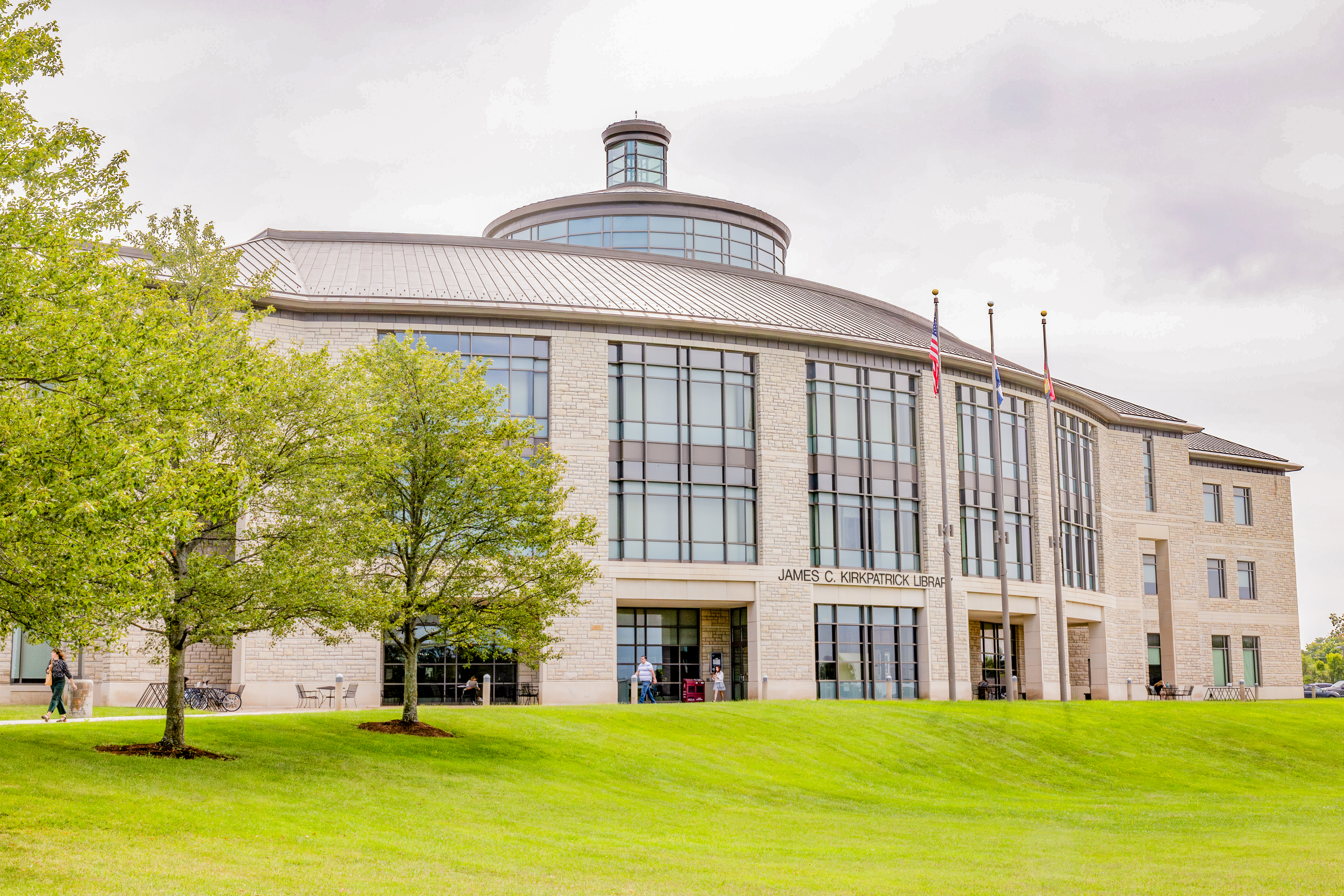 The James C. Kirkpatrick Library is seen. Several green leafy trees are on the left, and the bottom third of the photo is sloping green lawn in front of the library. A few people are walking on the sidewalk in front of the library. It is an overcast day.
