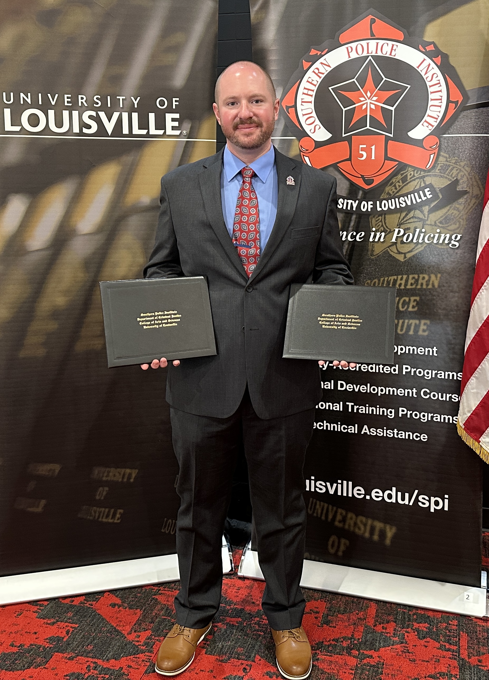Joe Jennings at SPI A person is wearing a dark-colored suit and holding two diploma/award covers. The person is looking at the camera and standing in front of two banners advertising the Southern Police Institute.