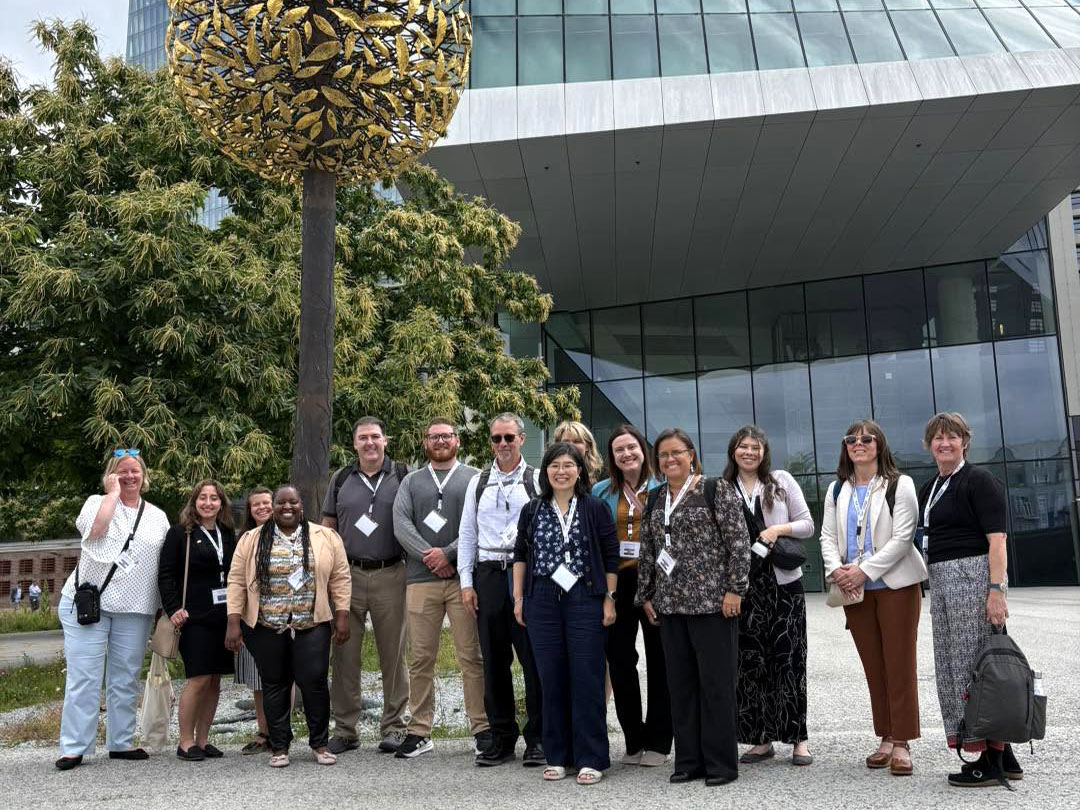 TOP group in Germany Fourteen people stand together in front of the European Central Bank, a glass and metal building. A tree and a tall metal sculpture are also seen behind t hem.