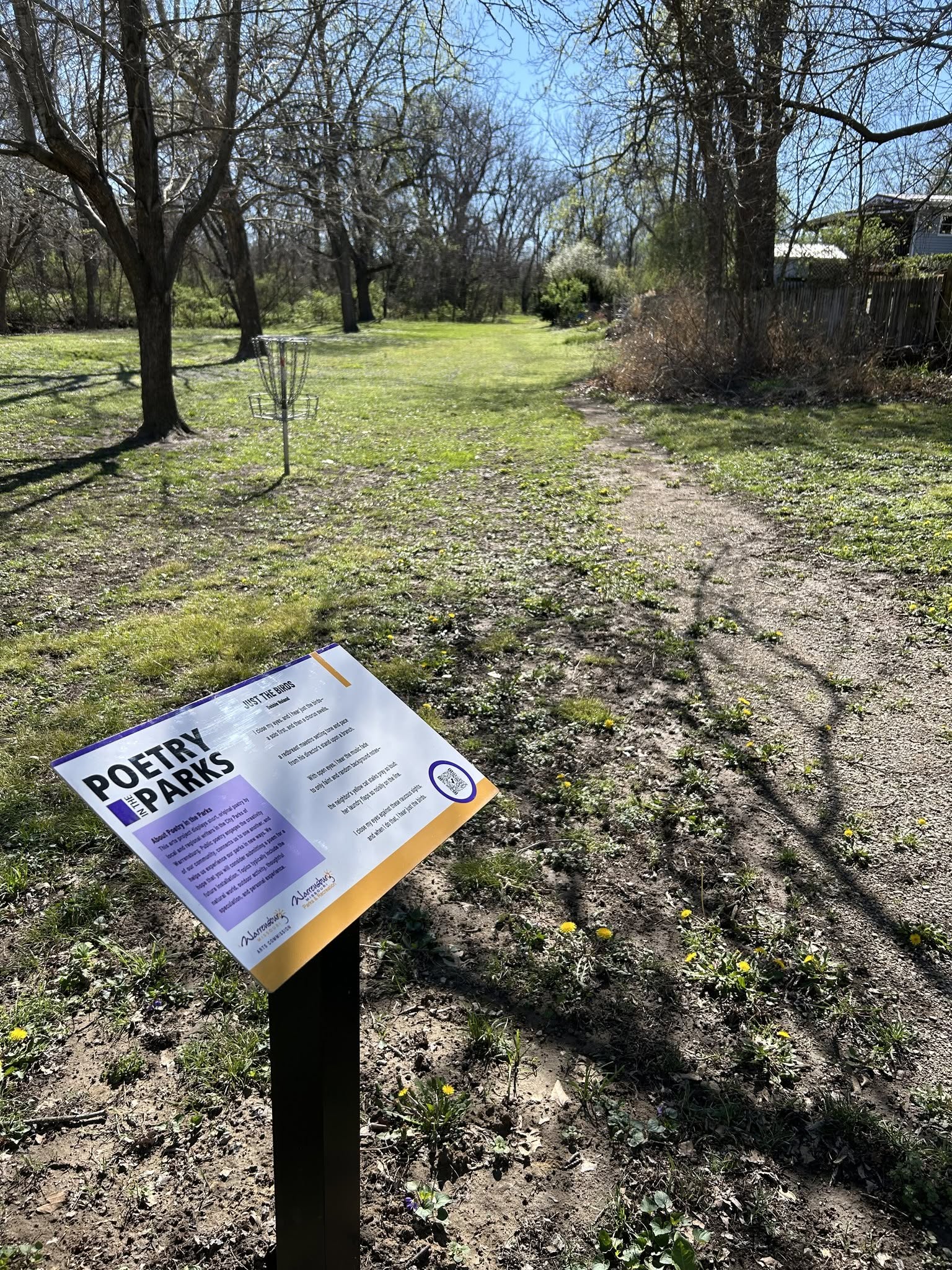 Poetry in the Parks A sign for the Poetry in the Parks project is seen in a grassy area of a park near a dirt trail.