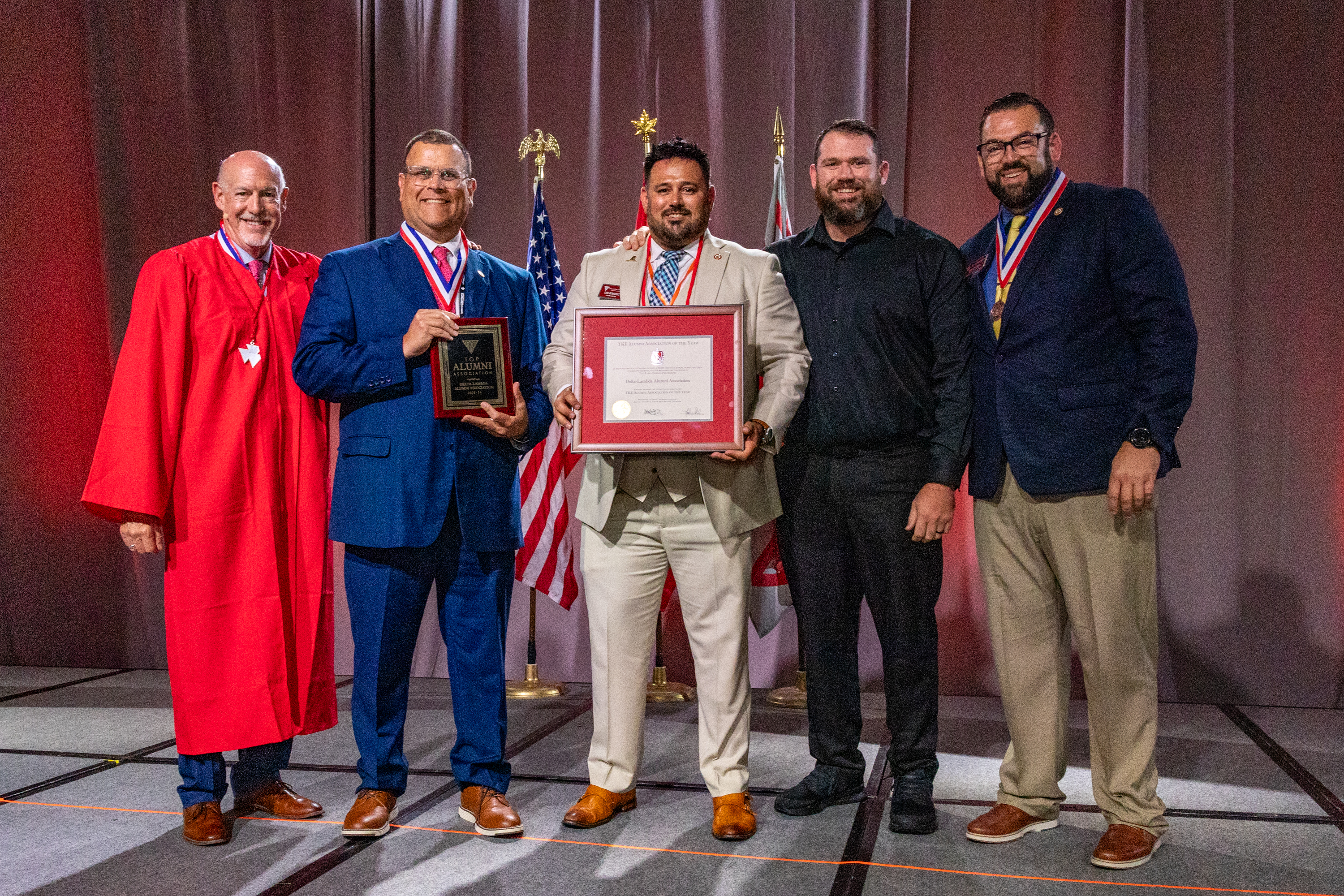 Tau Kappa Epsilon Fraternity awards Five people wearing suits or business clothing stand next to each other smiling on a stage. Two of them are each holding an award plaque.