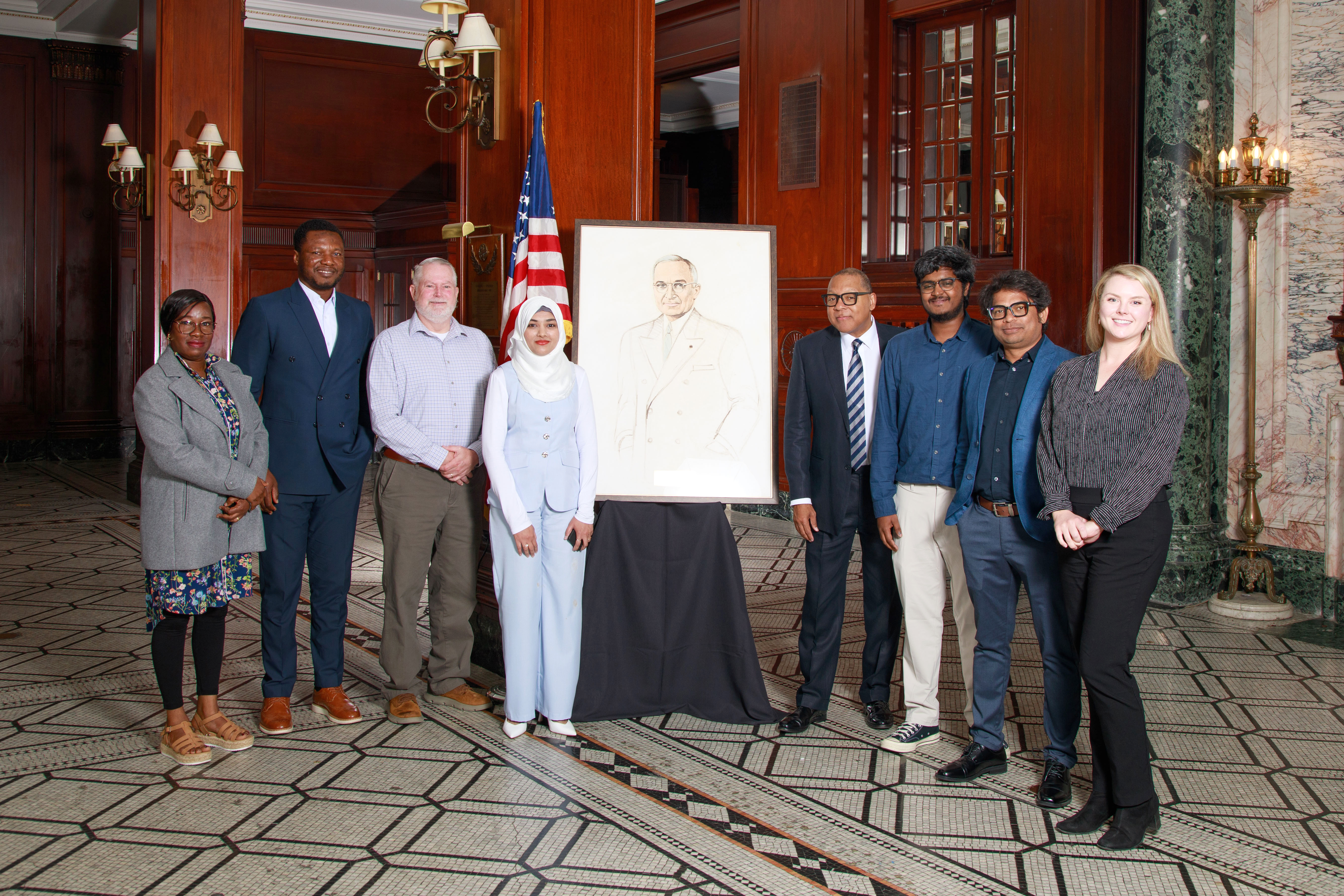 Good Neighbor Award Luncheon Eight people are standing in a line with an American flag and a painting of President Harry S. Truman in the middle. They are in an upscale hotel with ornate tile floor, marble pillars, wall sconces and dark wood walls.
