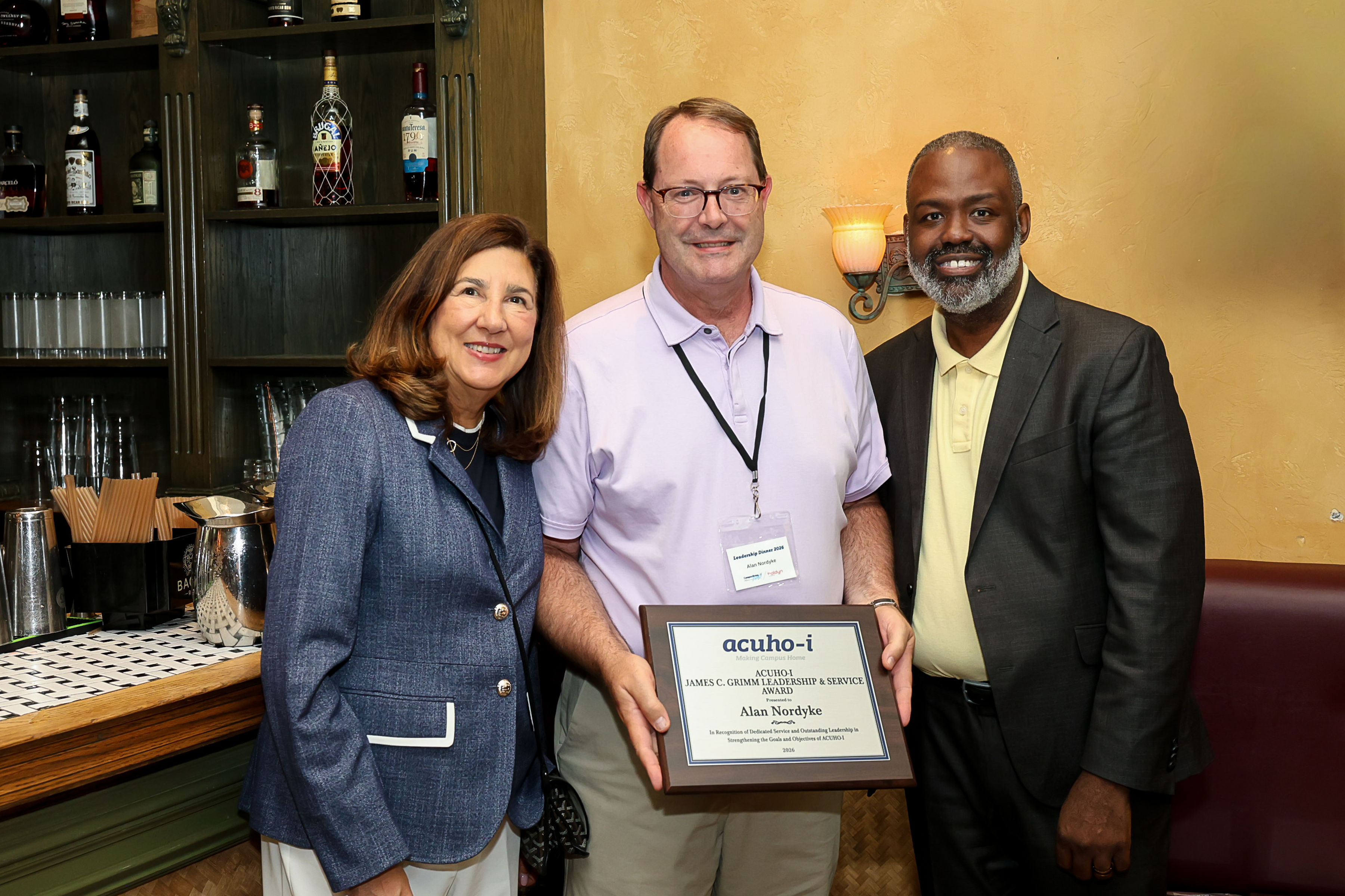 Nordyke award Three people stand next to each other in front of a bar in a restaurant. The award winner, Alan Nordyke, is in the middle and wearing a lavender polo and khaki pants and holding an award plaque. The person on the left is wearing a blue blazer and white pants, and the person on the right is wearing a dark gray suit jacket and pale yellow polo.