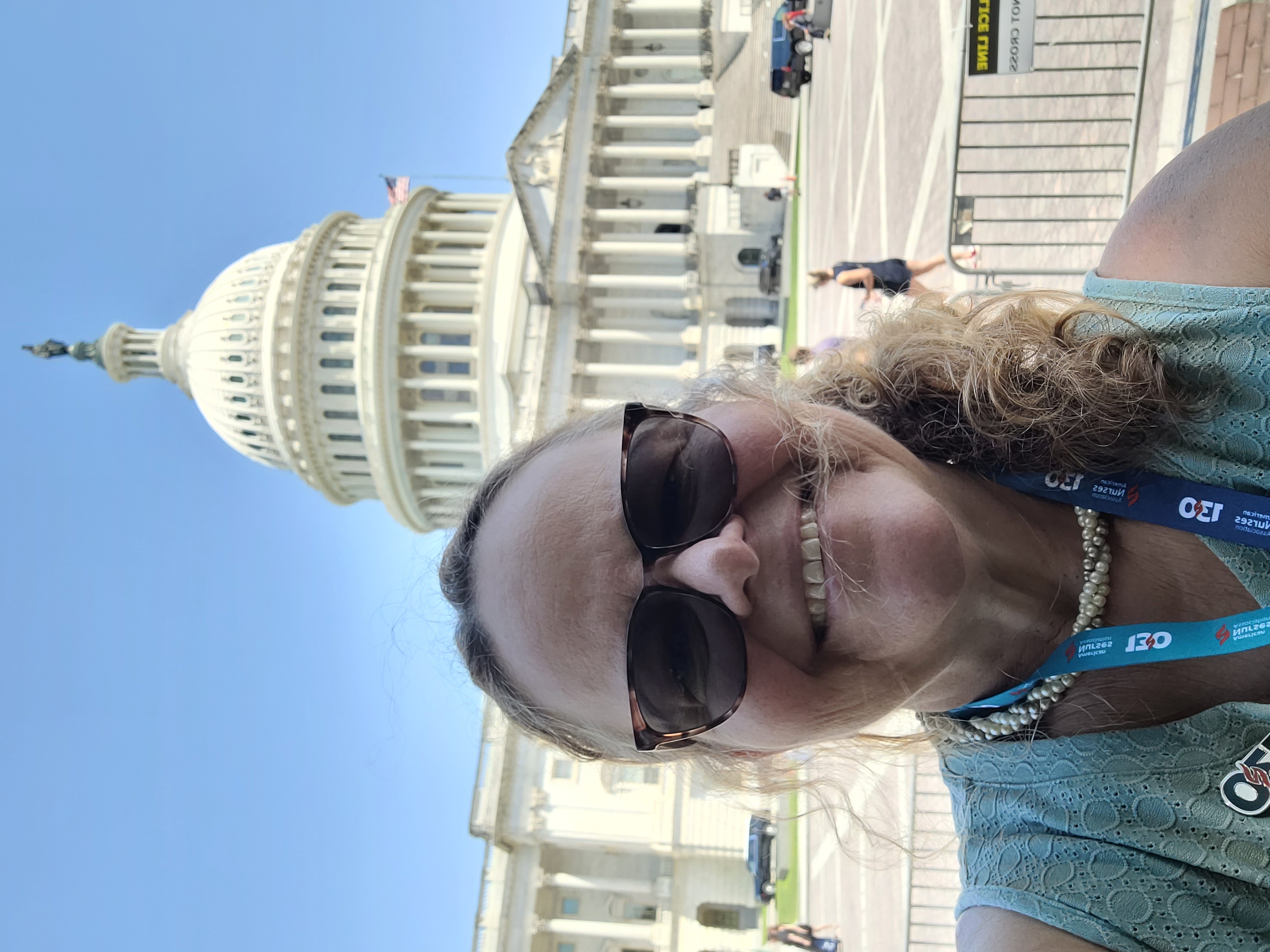 Janice Putnam A person is seen in a selfie photo in front of the U.S. Capitol building. The person is wearing sunglasses and a teal tank top, along with a necklace and lanyard for the conference.