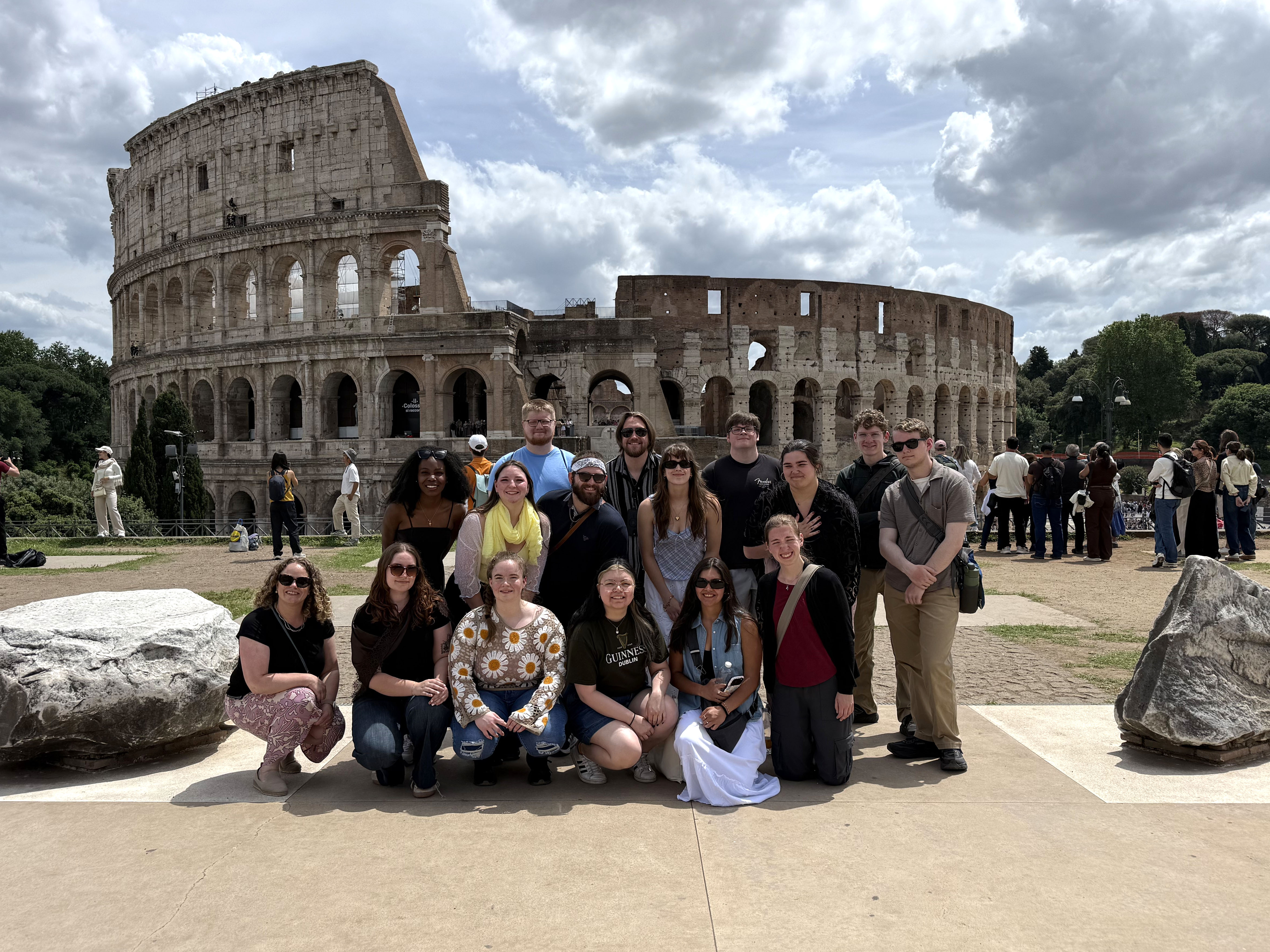 Honors College students and faculty in Rome Three rows of people, a total of 16 people, are smiling and posing for a photo in front of the Coliseum in Rome, Italy. Other tourists can be seen waiting in line behind them.