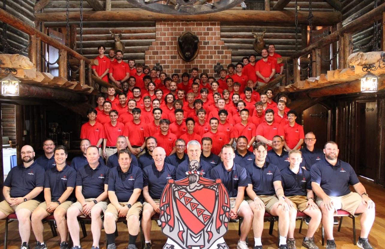 TKE Leadership Academy A group photo of facilitators and collegiate participants. The first two rows are facilitators wearing blue polos and khaki shorts or pants. Behind them are several rows of fraternity members wearing red polos. The Tau Kappa Epsilon Fraternity coat of arms is seen at the front of the photo. About 100 people are in the photo.