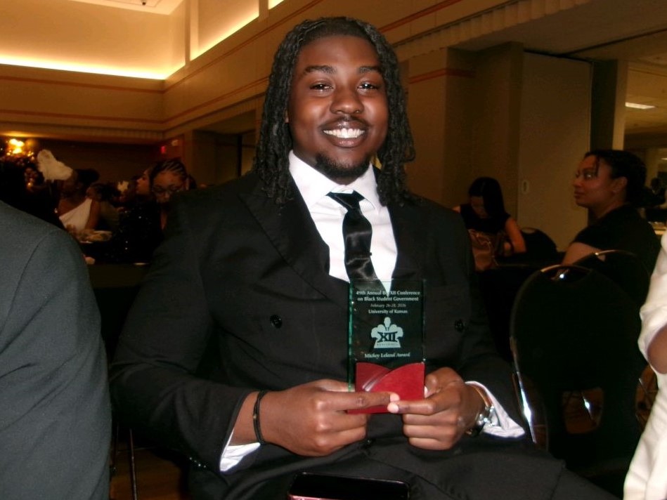 A person is sitting in a chair while smiling and holding an award plaque.