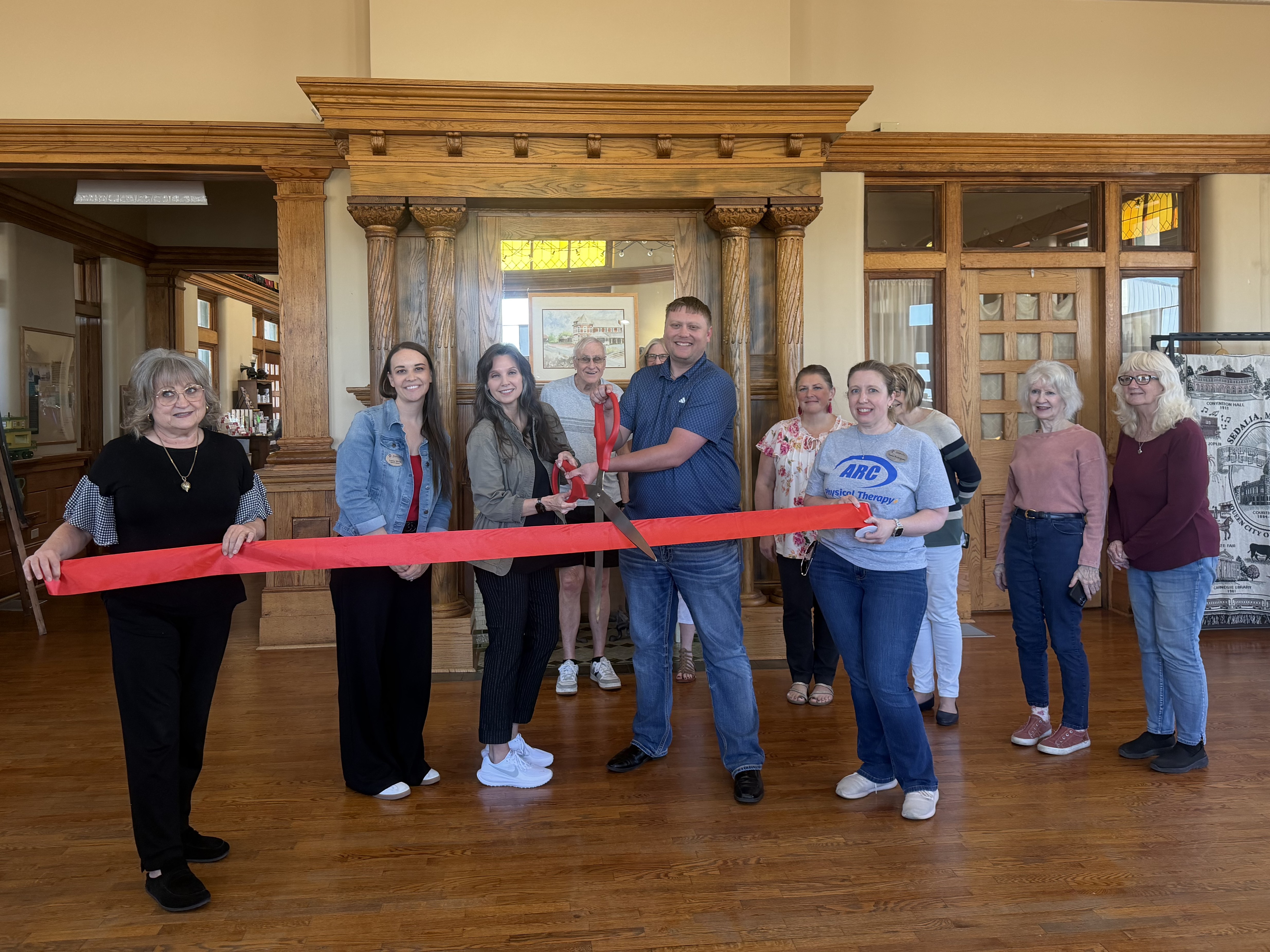 Two people are holding either side of a long red ribbon as a person in the middle starts to cut the ribbon with huge ceremonial scissors. Other people are standing around them.