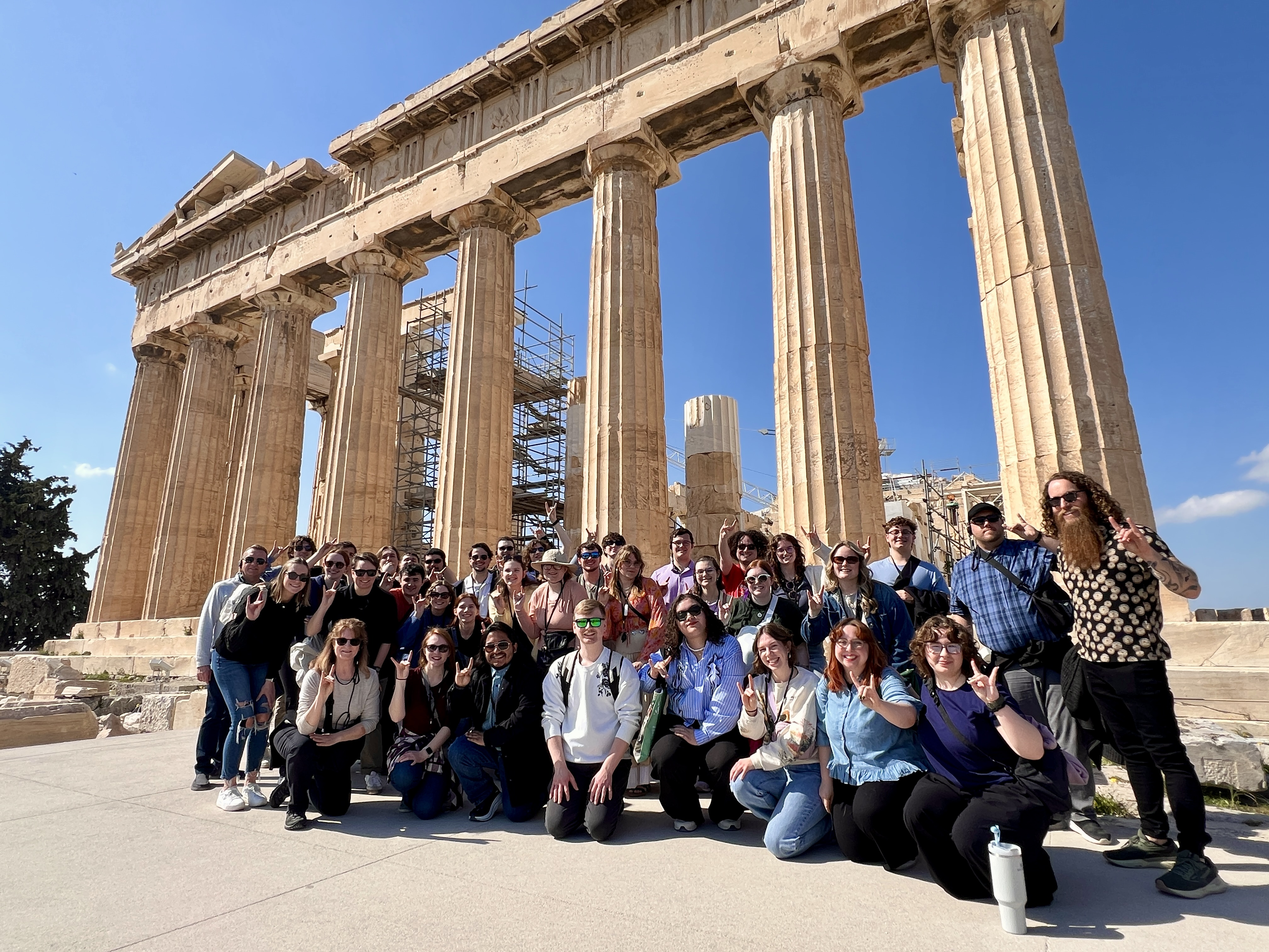 A large group of people stands in front of the ancient ruins of the Parthenon in Greece.