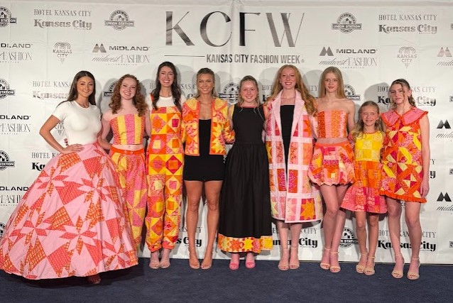 Nine people stand in a row with a white backdrop behind them that says Kansas City Fashion Week.