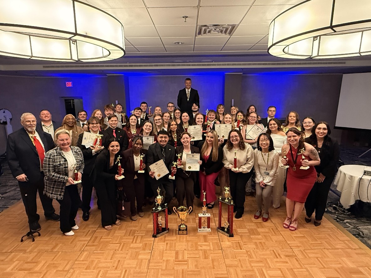 A large group of people stand together inside a room with a wood floor. Several people in the front row are holding trophies and certificates.