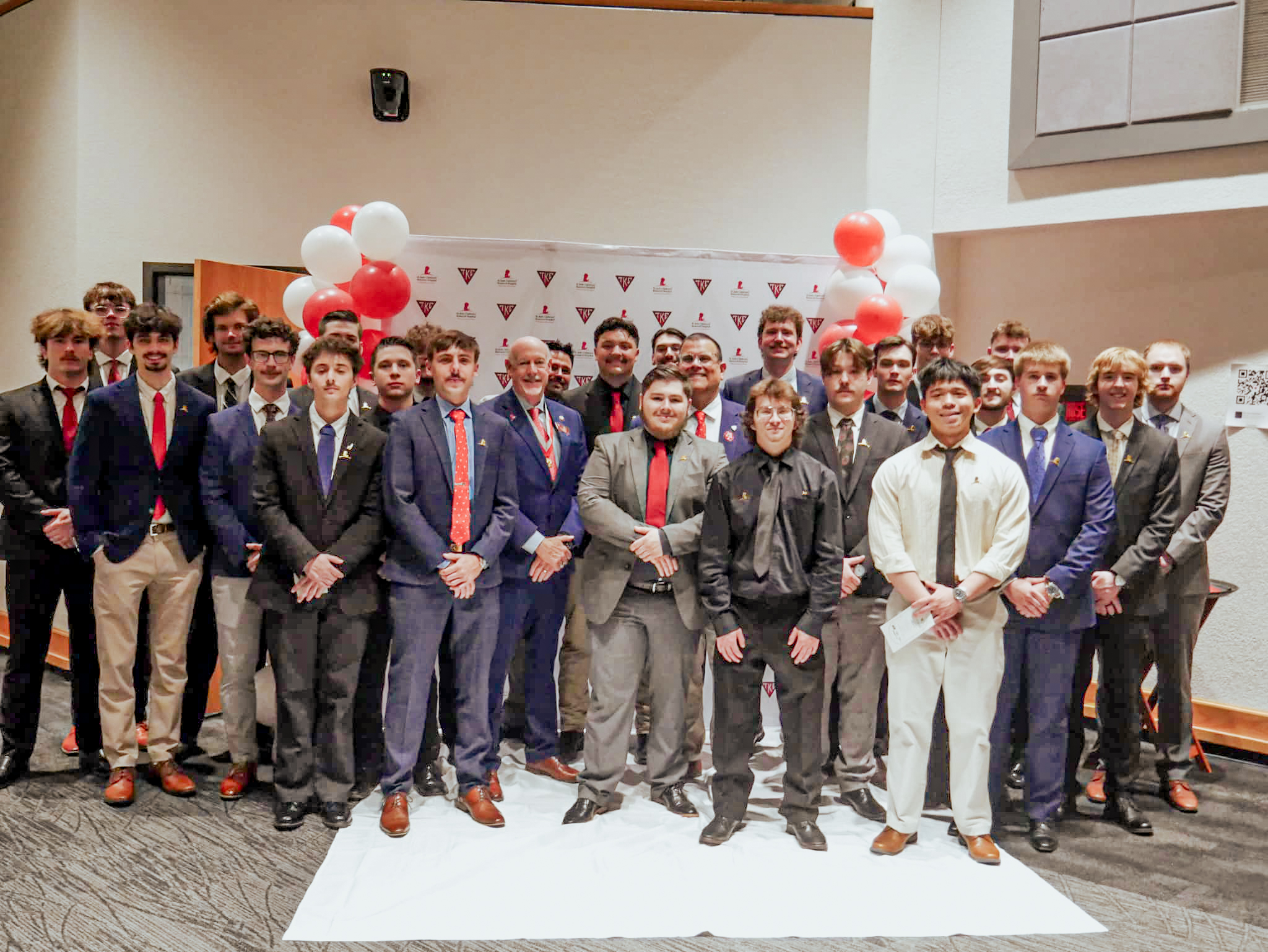 A group of people poses for a photo in the Elliott Student Union Ballroom. Red and white balloons and a white backdrop with the TKE Fraternity and St. Jude's Hospital logos are behind the group.