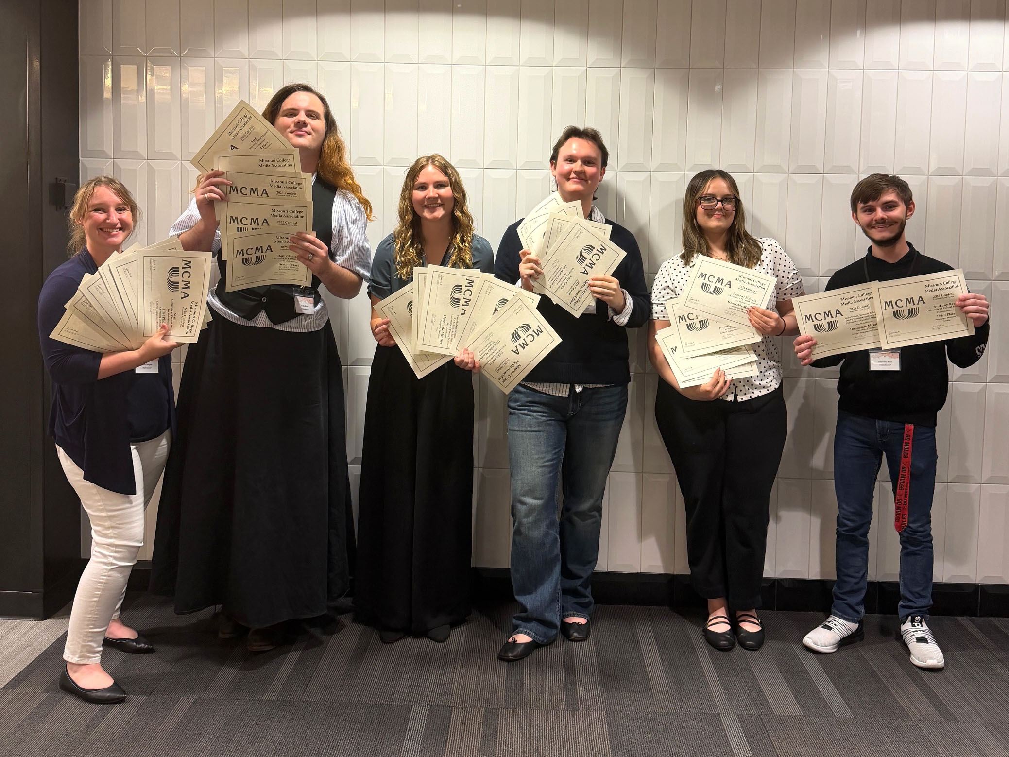 Six students stand next to each other in a line and in front of a white textured wall. Each person is holding multiple paper award certificates.
