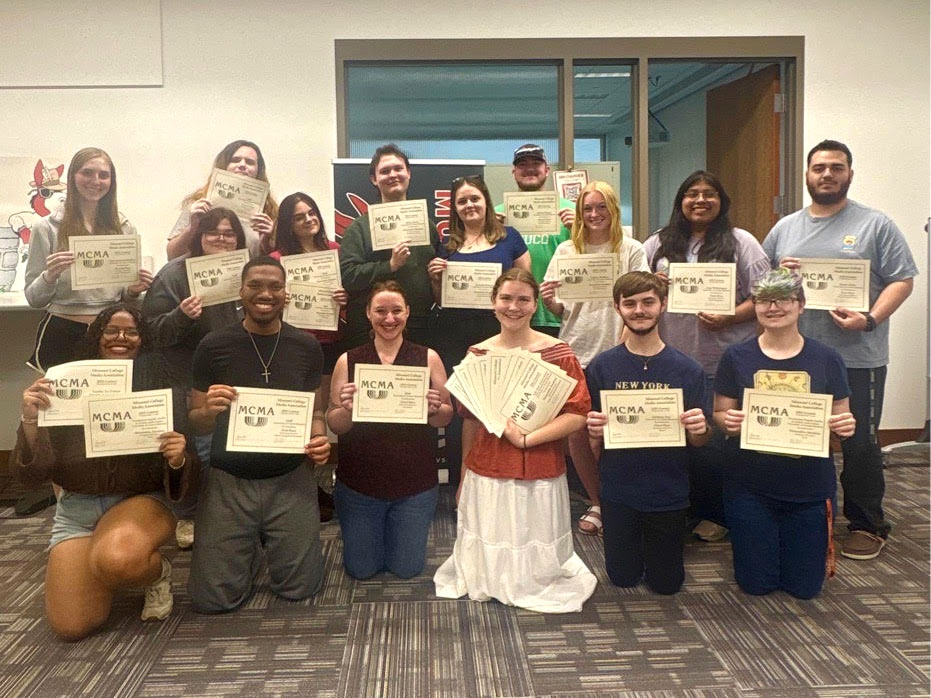 Sixteen students are posing for a group photo in the library in front of the Muleskinner newsroom. Six people are kneeling in the front row and the others are standing behind them. Everyone is holding one or multiple paper award certificates.