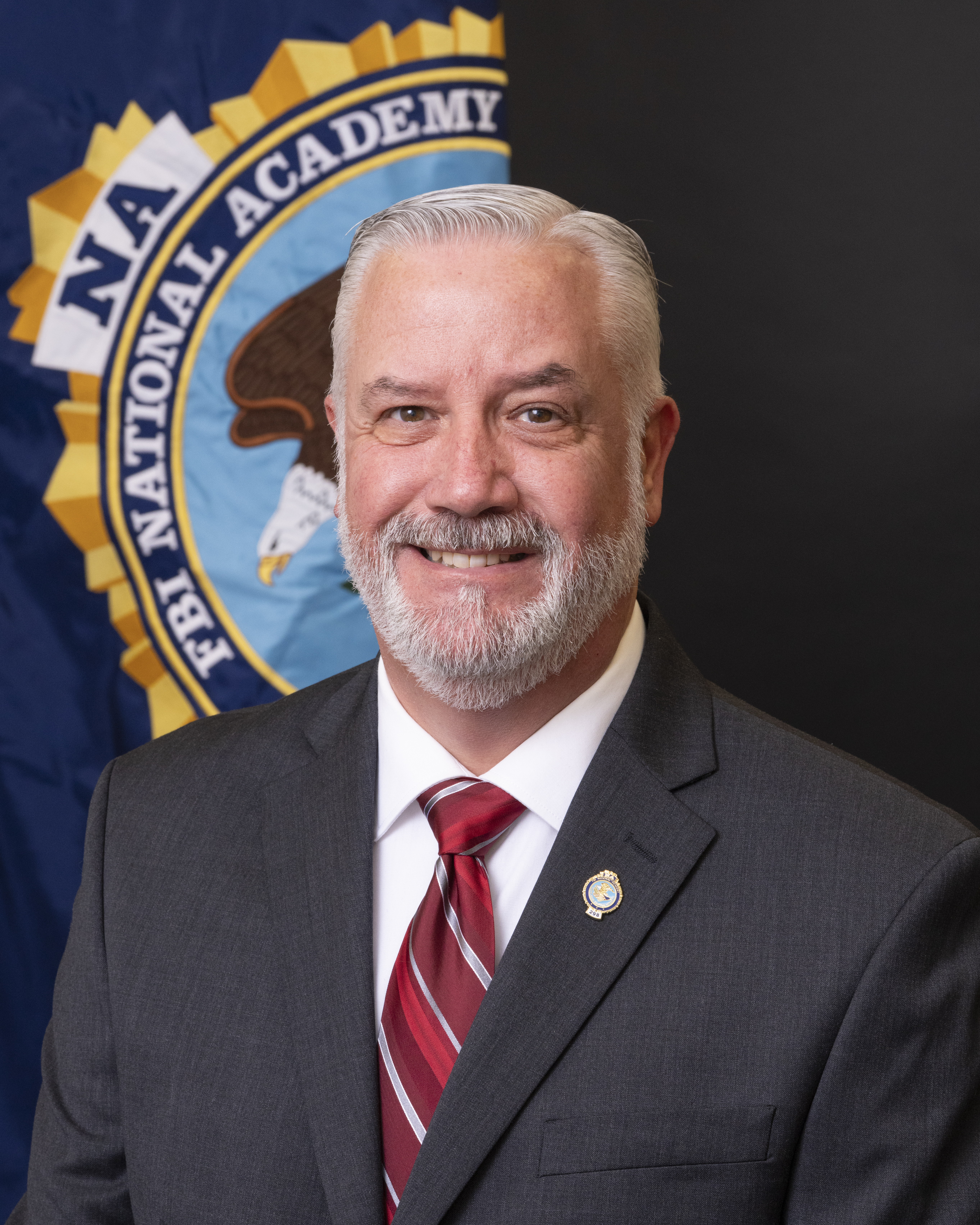 Capt. Mike Papasifakis A person is seen in a professional portrait from the shoulders up. He is wearing a dark gray suit with a white shirt and red striped tie. Behind him, the FBI National Academy seal is partially seen.