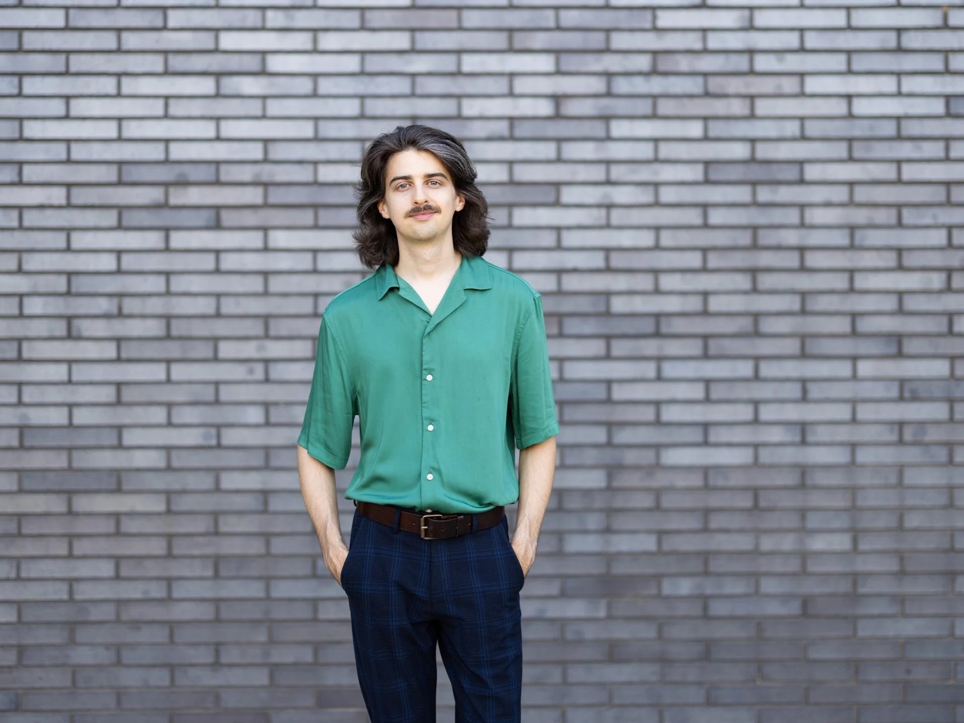 A person is standing in front of a gray brick wall. They have long, dark hair and a mustache and are wearing a green button-up collared shirt and navy plaid pants.