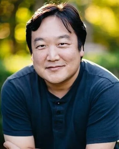 A man poses for a portrait and is seen from the shoulders up with greenery behind him