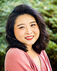A woman poses for a portrait and is seen from the shoulders up with greenery behind her