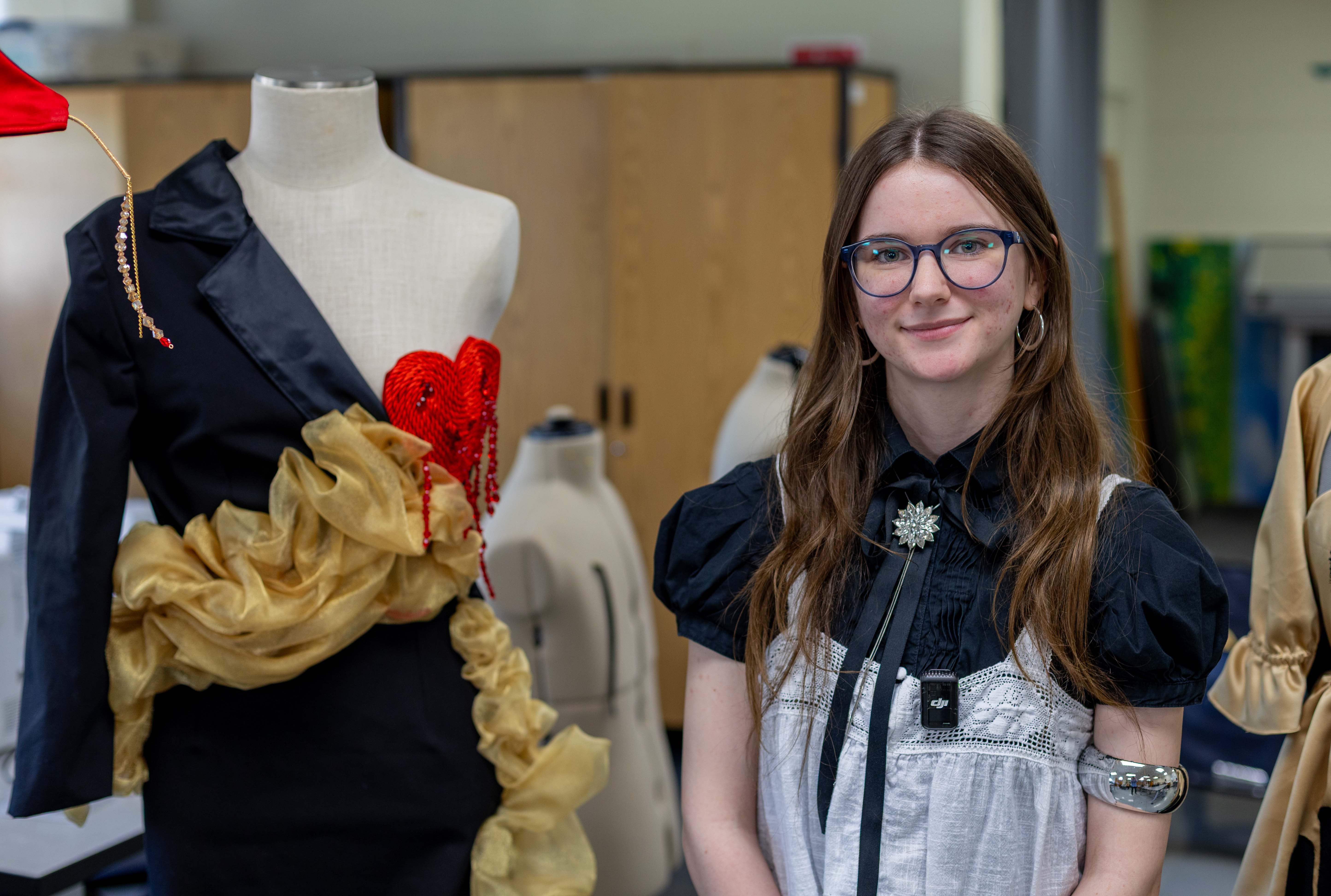 A person is wearing a black short-sleeve shirt under a white tank top dress and glasses. To the left is a dress on a mannequin and behind the person are several bare mannequins.