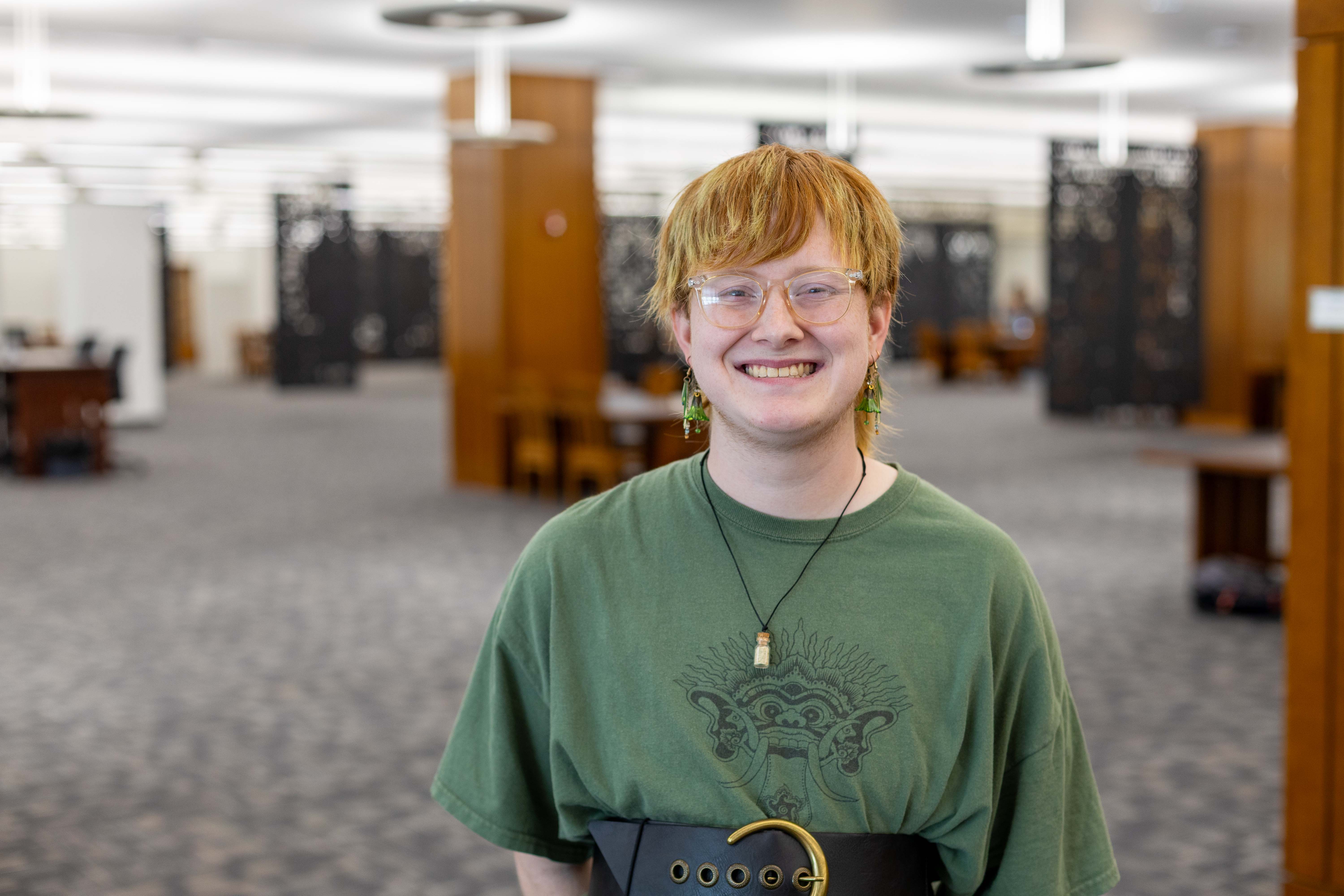 A person is wearing glasses, a green T-shirt, a necklace and green earrings while standing in a library. The carpet is gray and wooden worktables and other fixtures can be seen in the background.